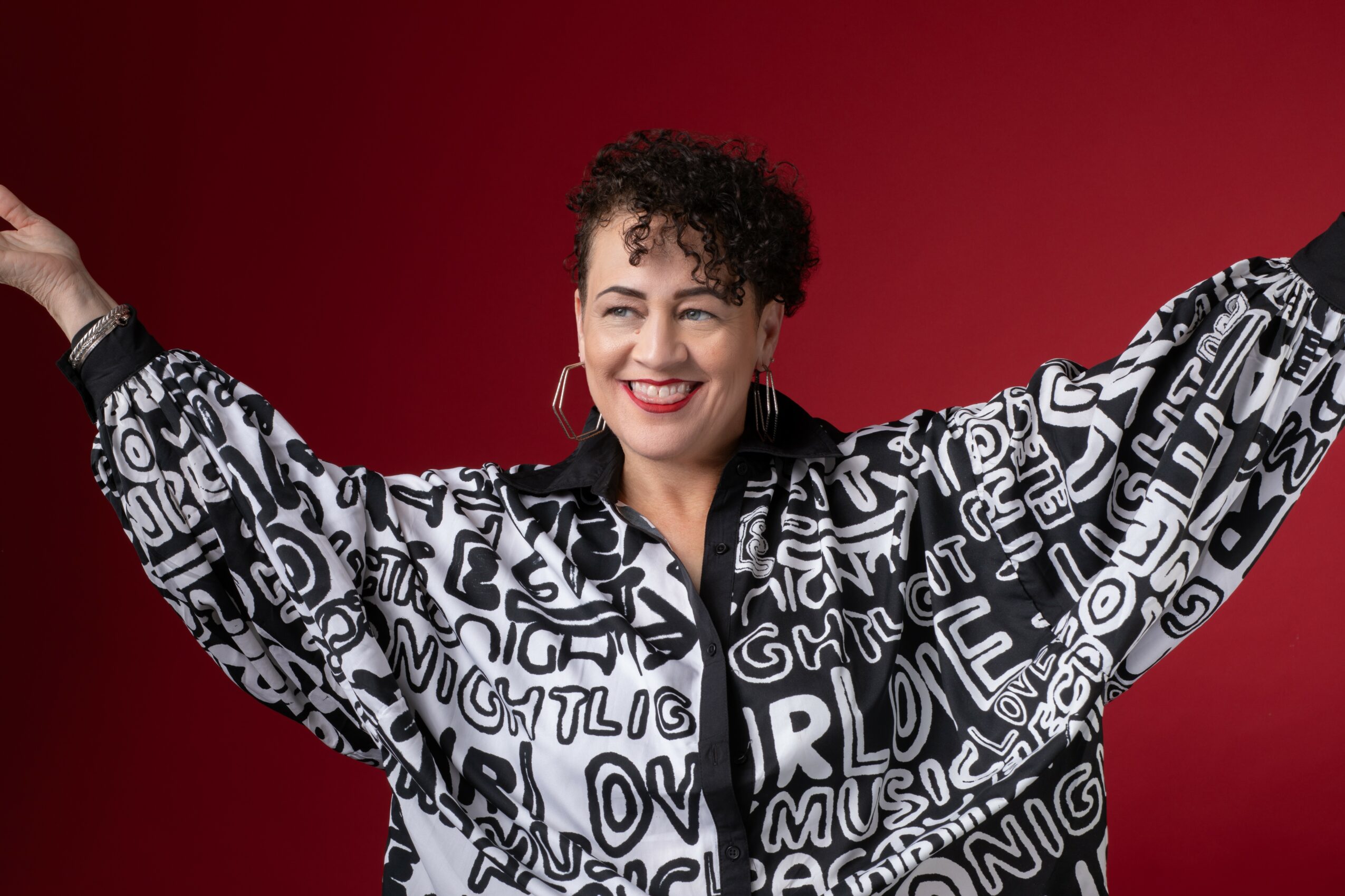 Woman with short curly hair smiling, wearing a black and white patterned shirt with text, against a red background.