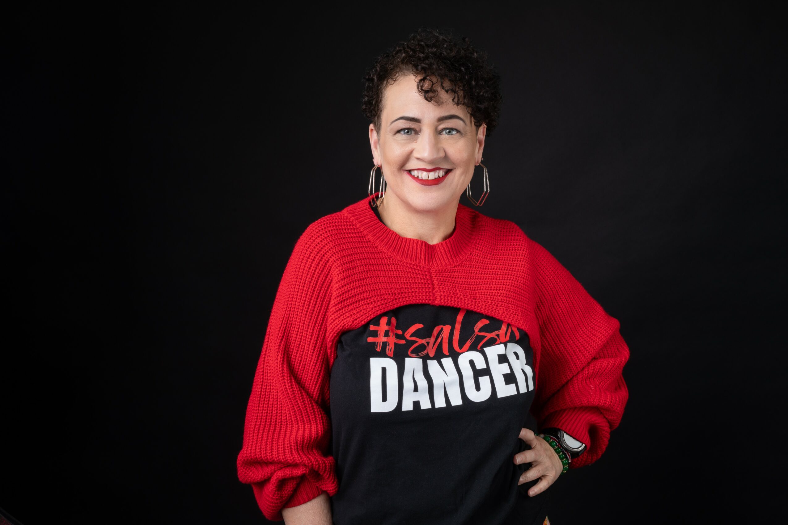 Woman with short curly hair smiling, wearing a red and black sweater with text, against a dark background.