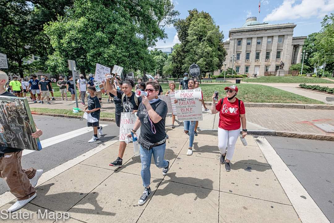 Group of people participating in a protest march on a city street, holding signs and banners, with a large building and trees in the background.