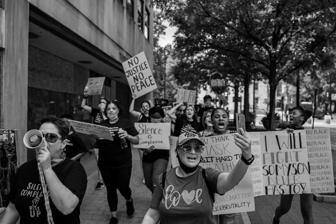 Group of people protesting with signs and megaphone outdoors, trees and buildings in background.