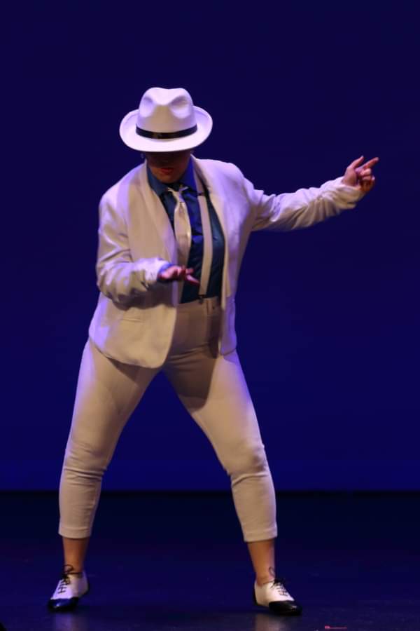 Person in white suit and hat dancing on stage with dark background.
