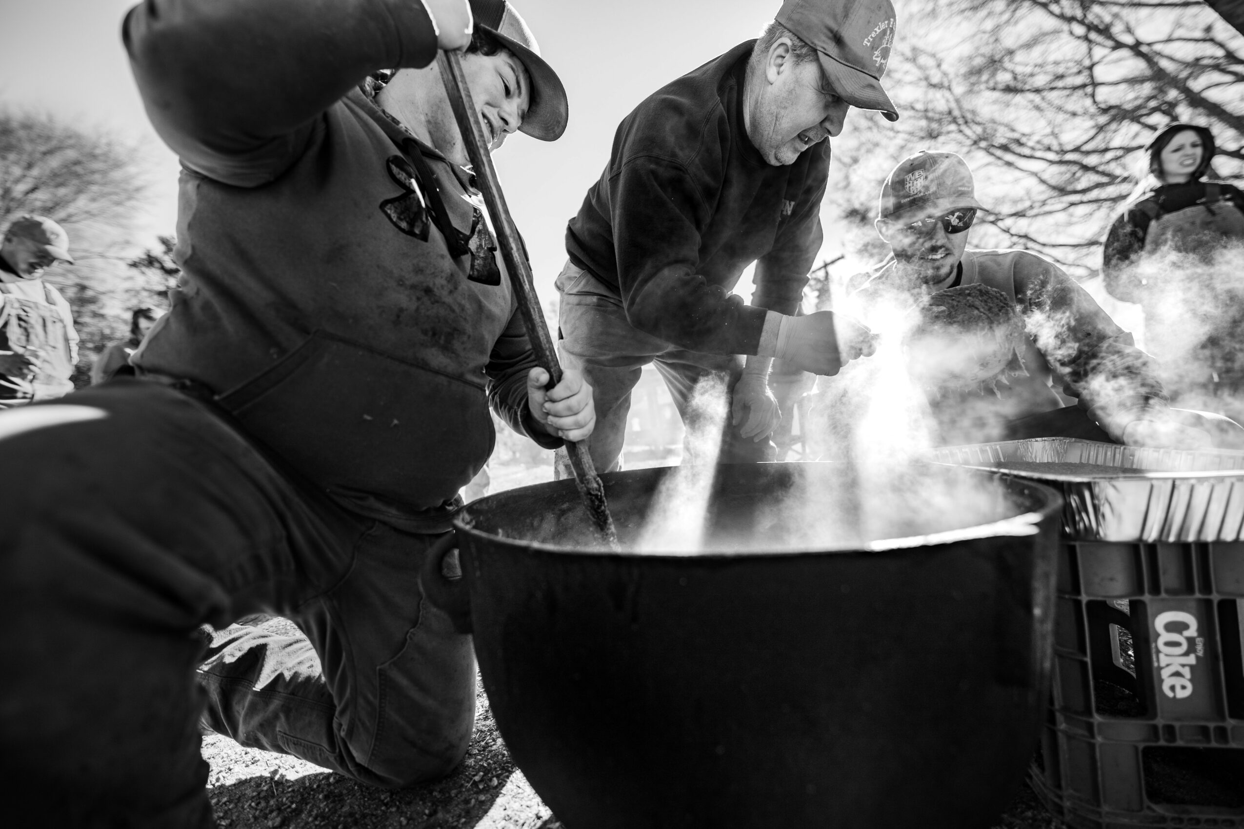 People gathered outdoors around a large steaming pot, with trees and other individuals in the background, in black and white.