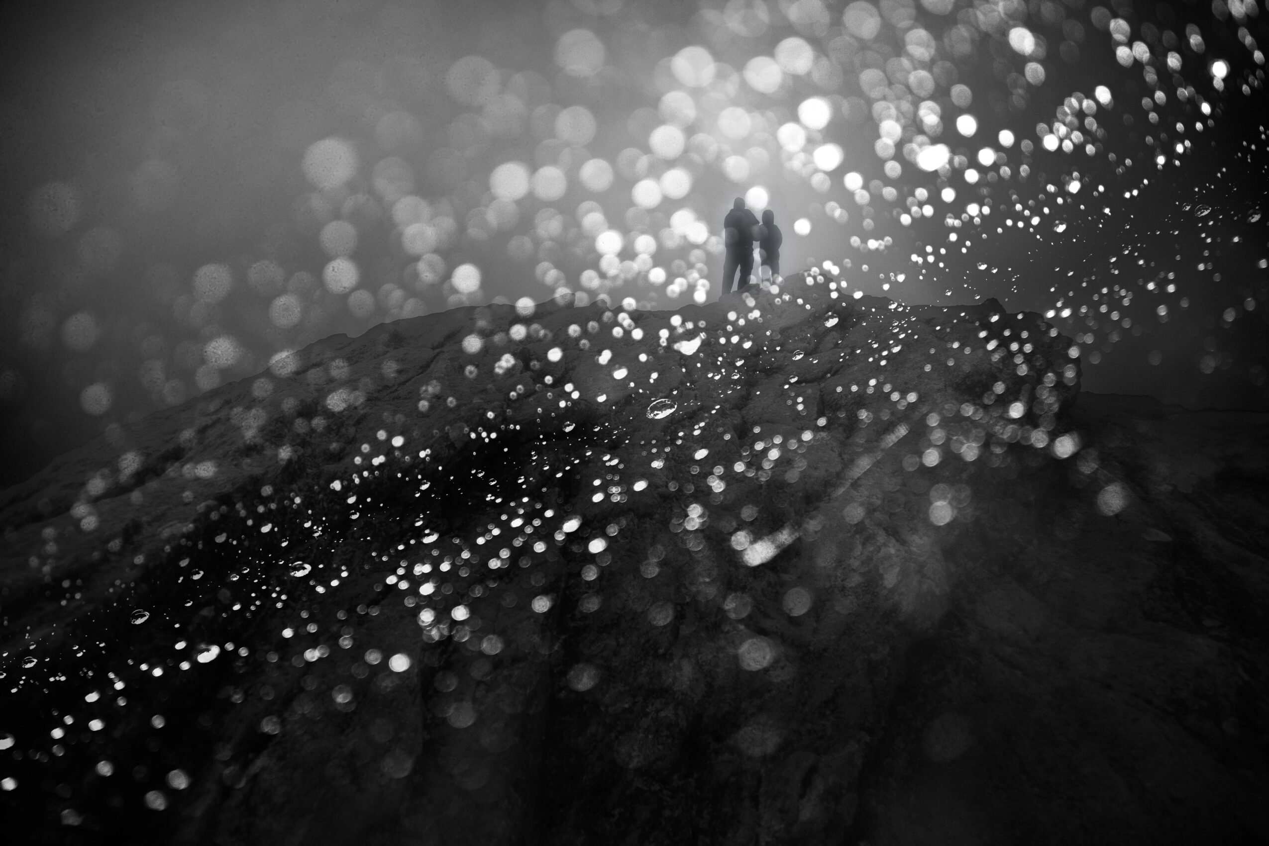 Two people standing on a dark, wet surface with bright, out-of-focus light spots in the background.