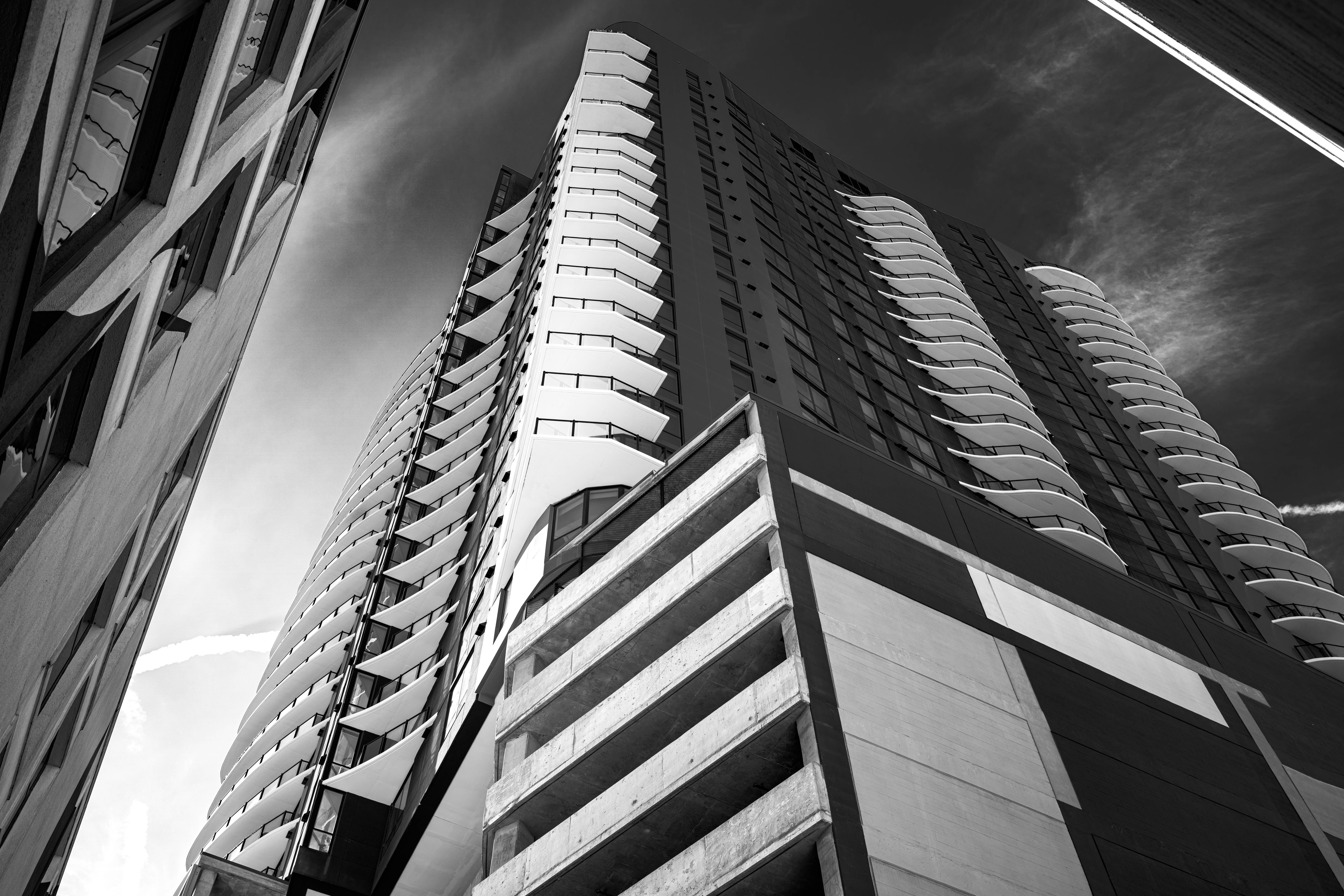 Tall modern building with curved balconies, viewed from below, against a cloudy sky.
