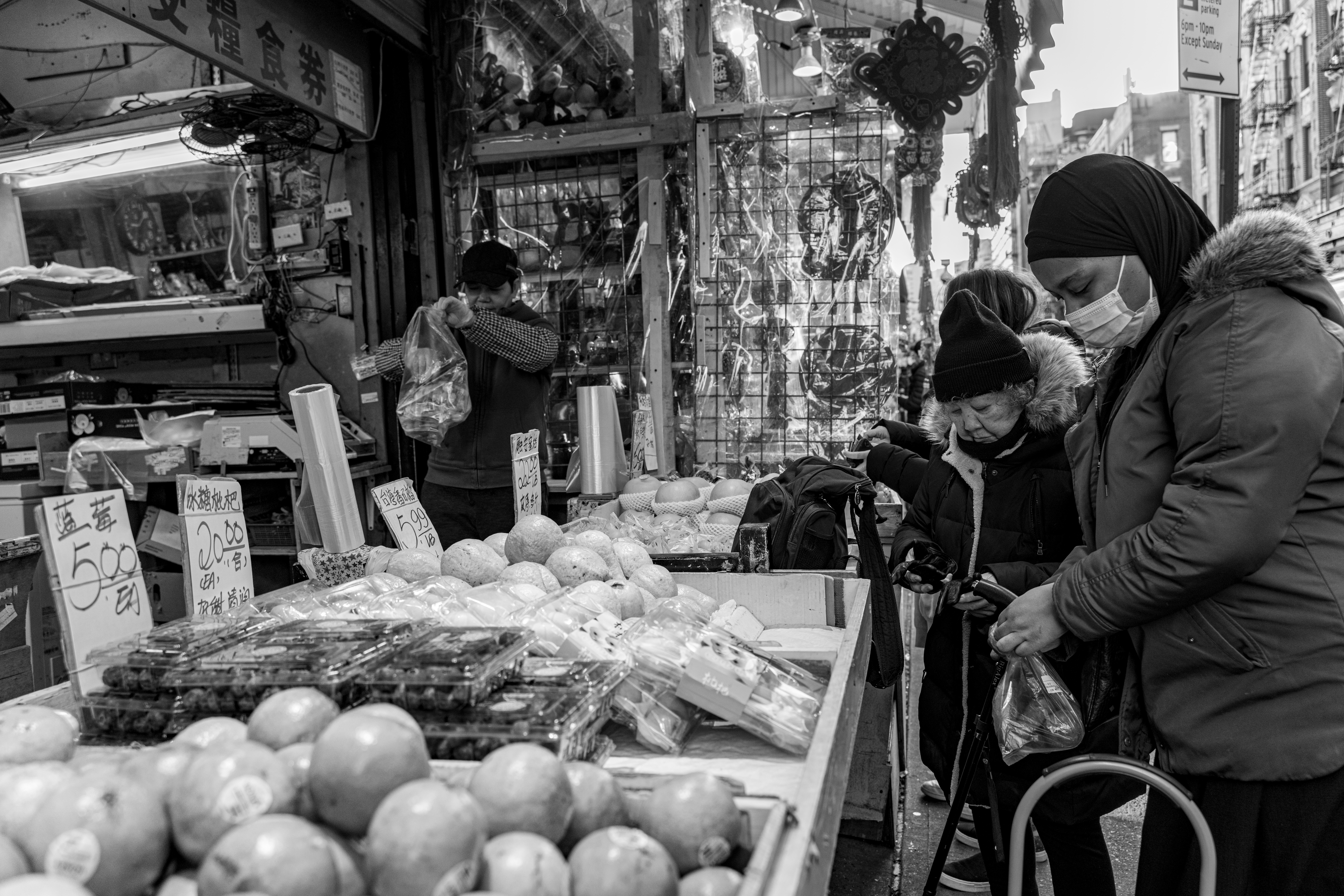 People shopping at a market stall with fruits and vegetables, wearing masks, in an urban setting, black and white photo.