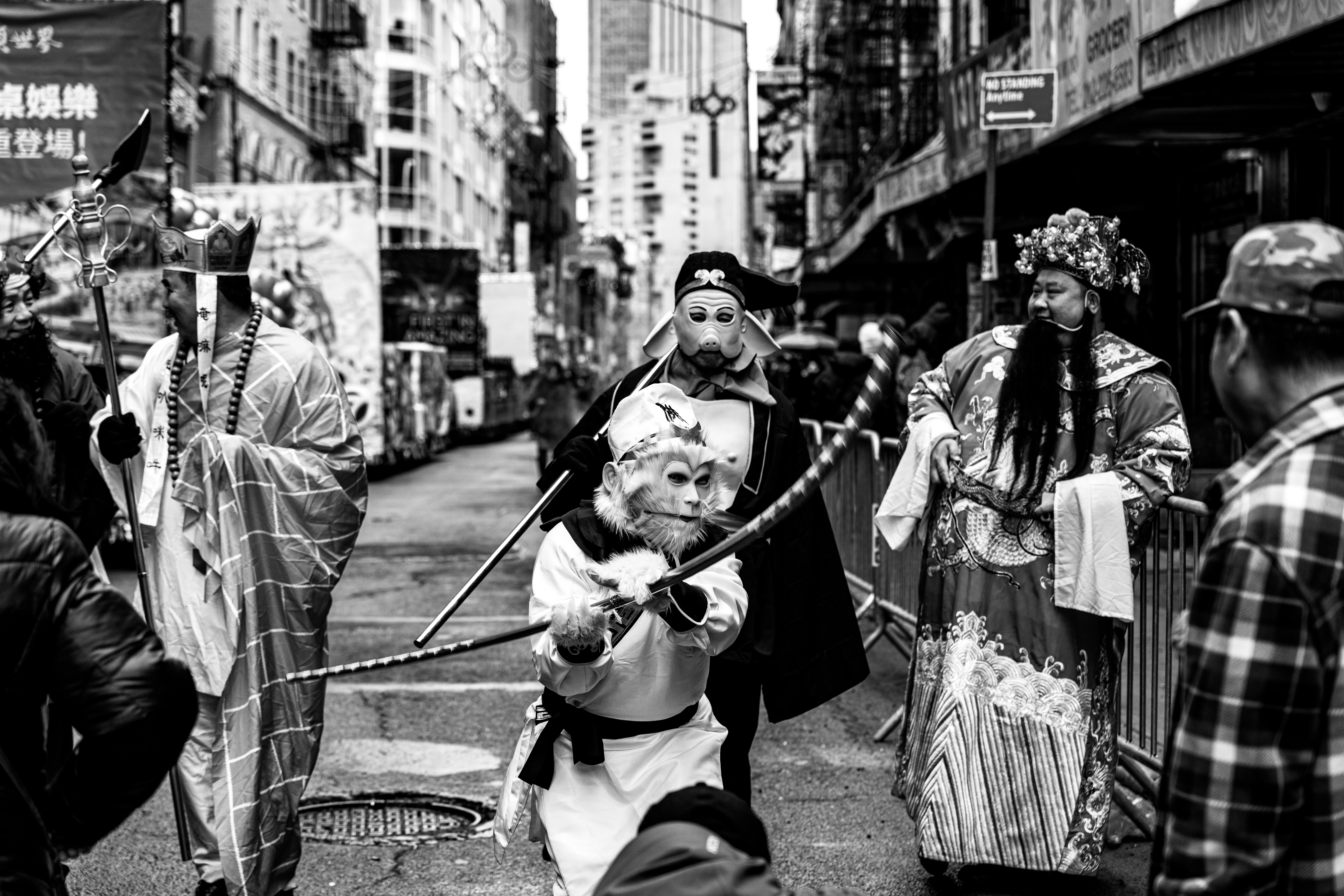 People in traditional costumes and masks gather on a city street with buildings and signs in the background.
