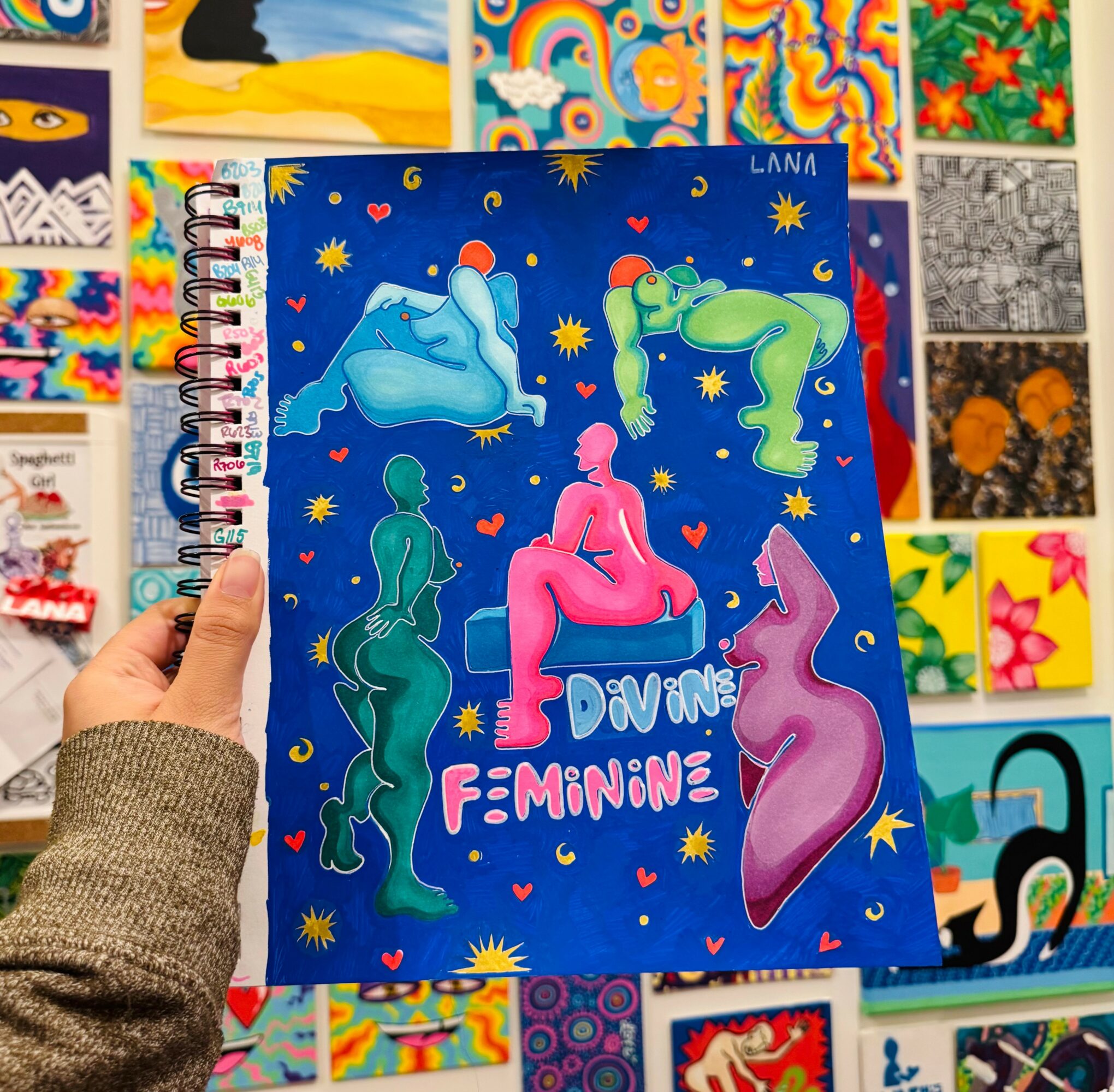 Hand holding a colorful notebook with abstract human figures and text, surrounded by vibrant artwork on a wall.