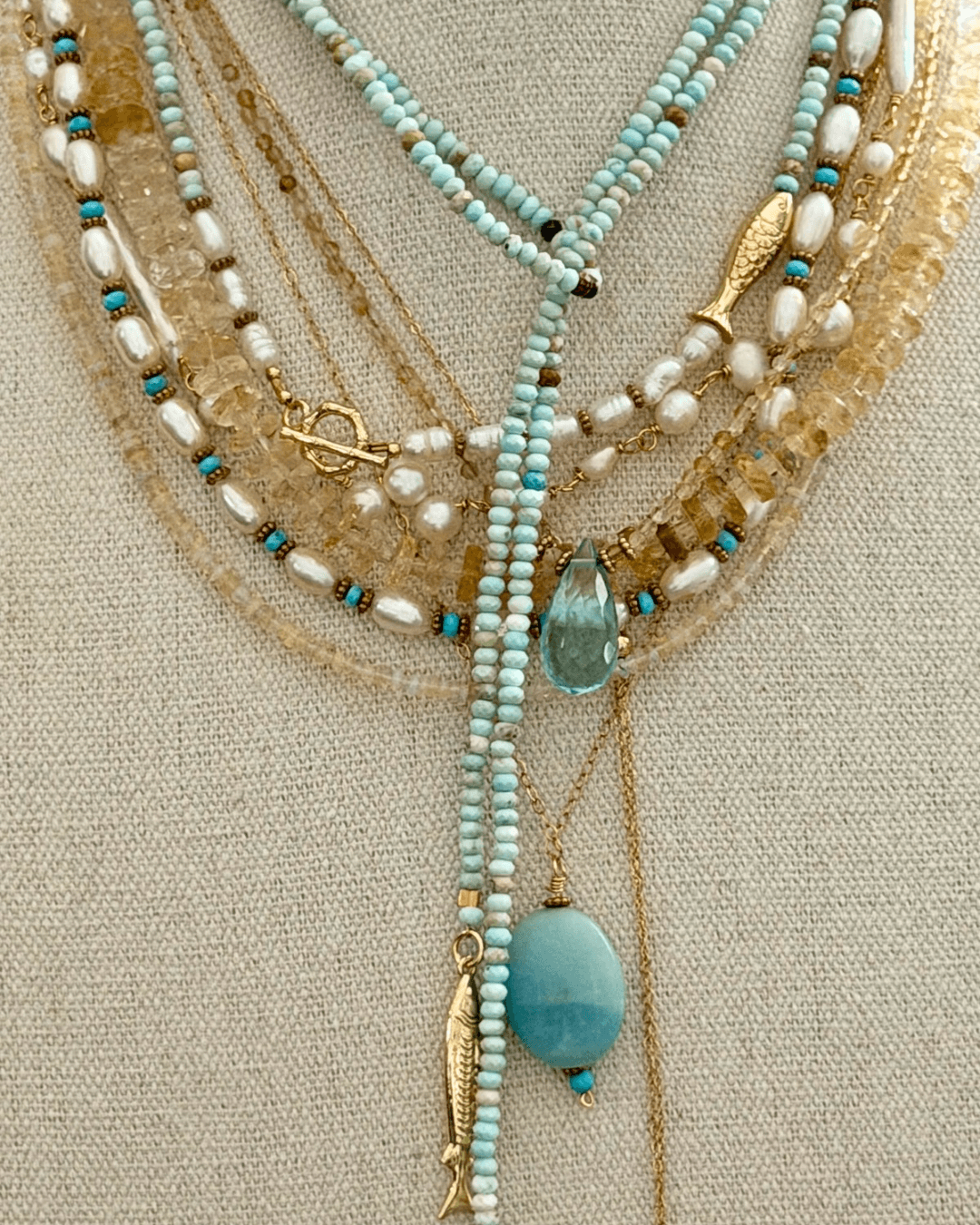 Multiple layered necklaces with beads, gold, and a large blue pendant displayed on a fabric background.