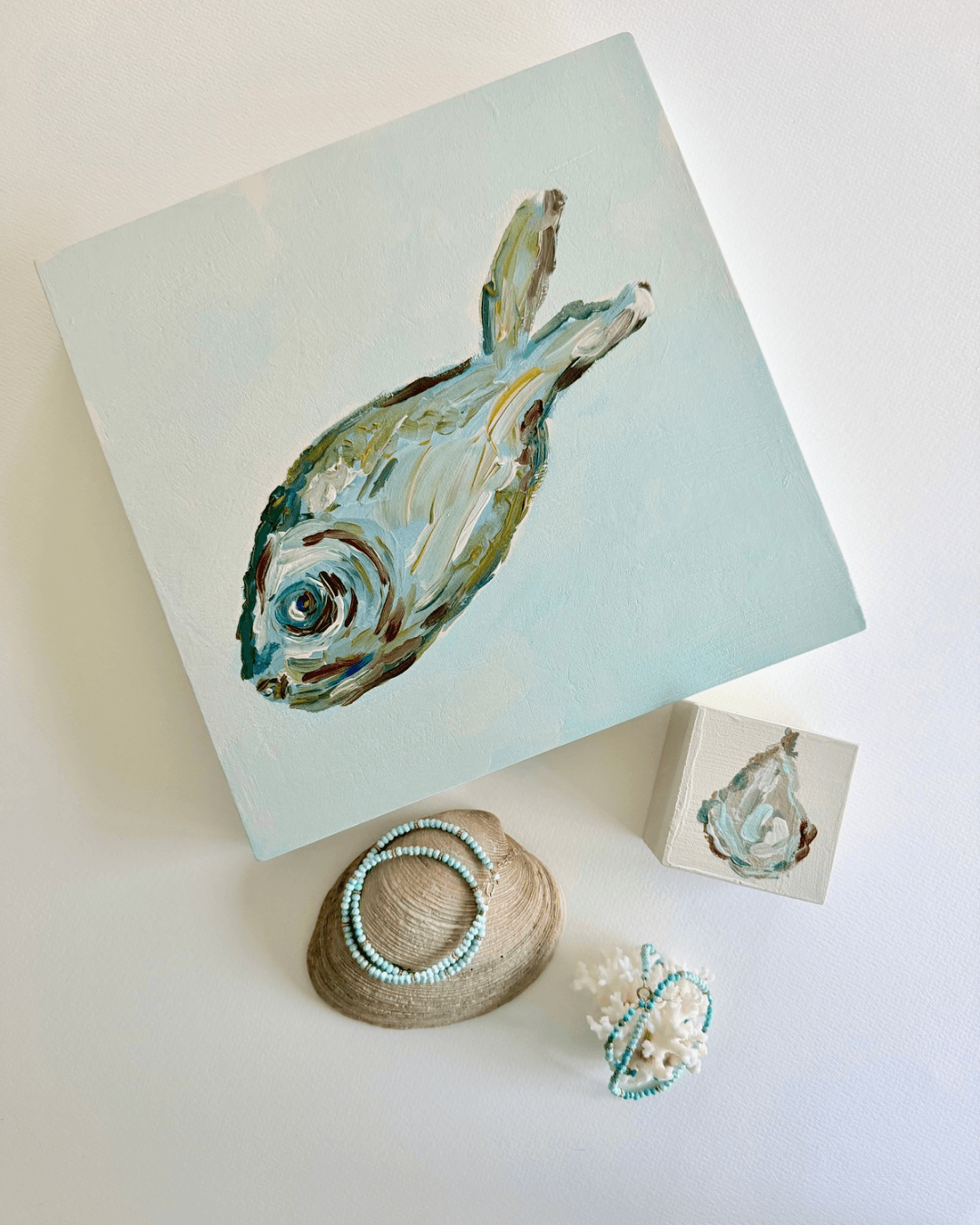 Painting of a fish on a light background, with a small matching artwork, shells, and jewelry nearby.