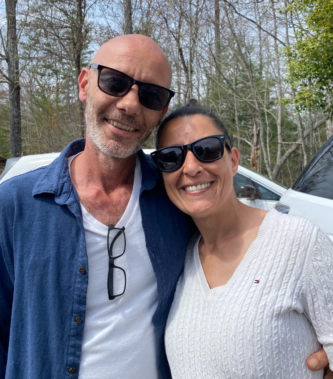 Two people smiling outdoors, wearing sunglasses, standing close together with trees in the background.