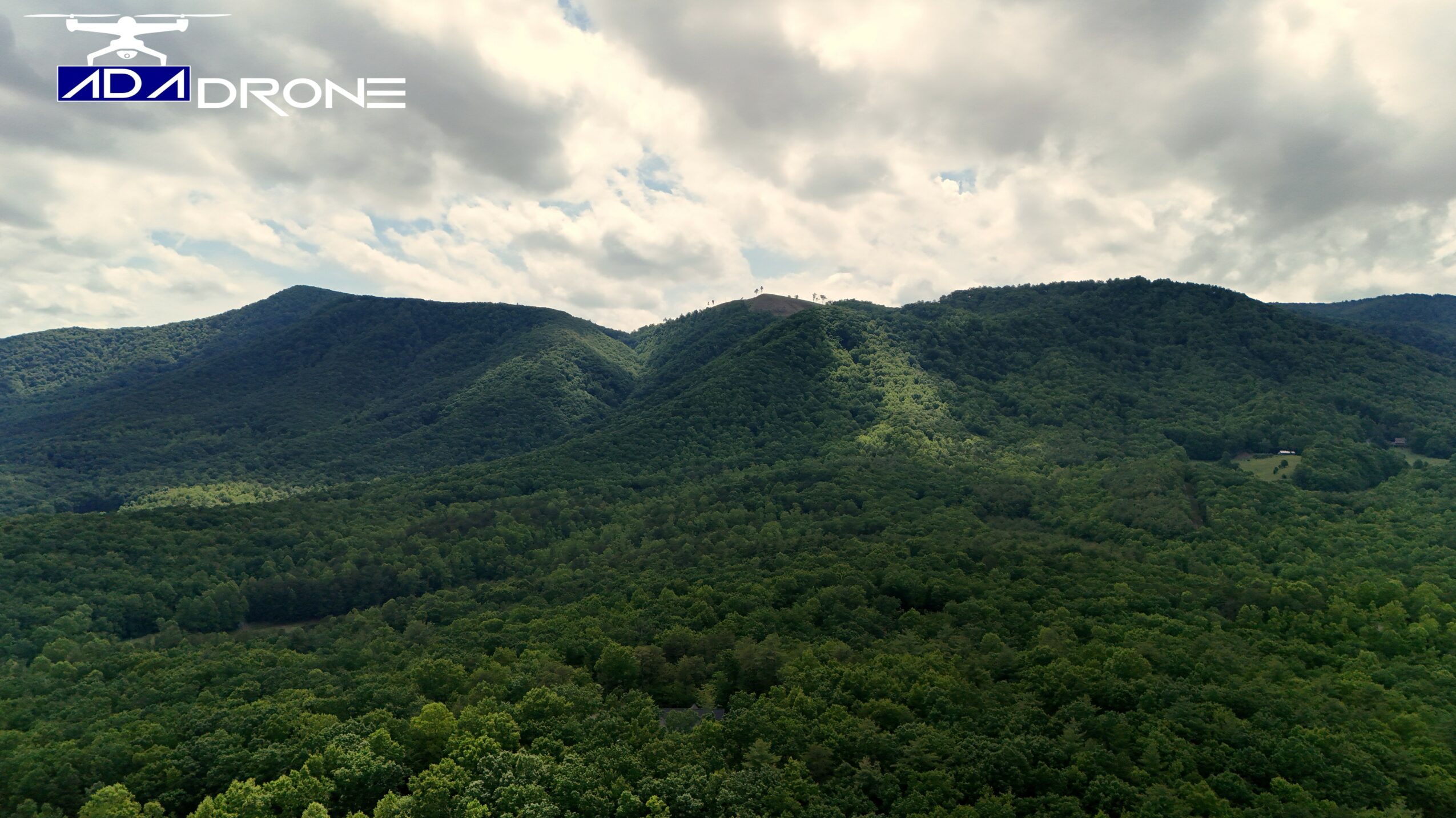 Landscape of green forested mountains under a partly cloudy sky, with a logo in the top left corner.