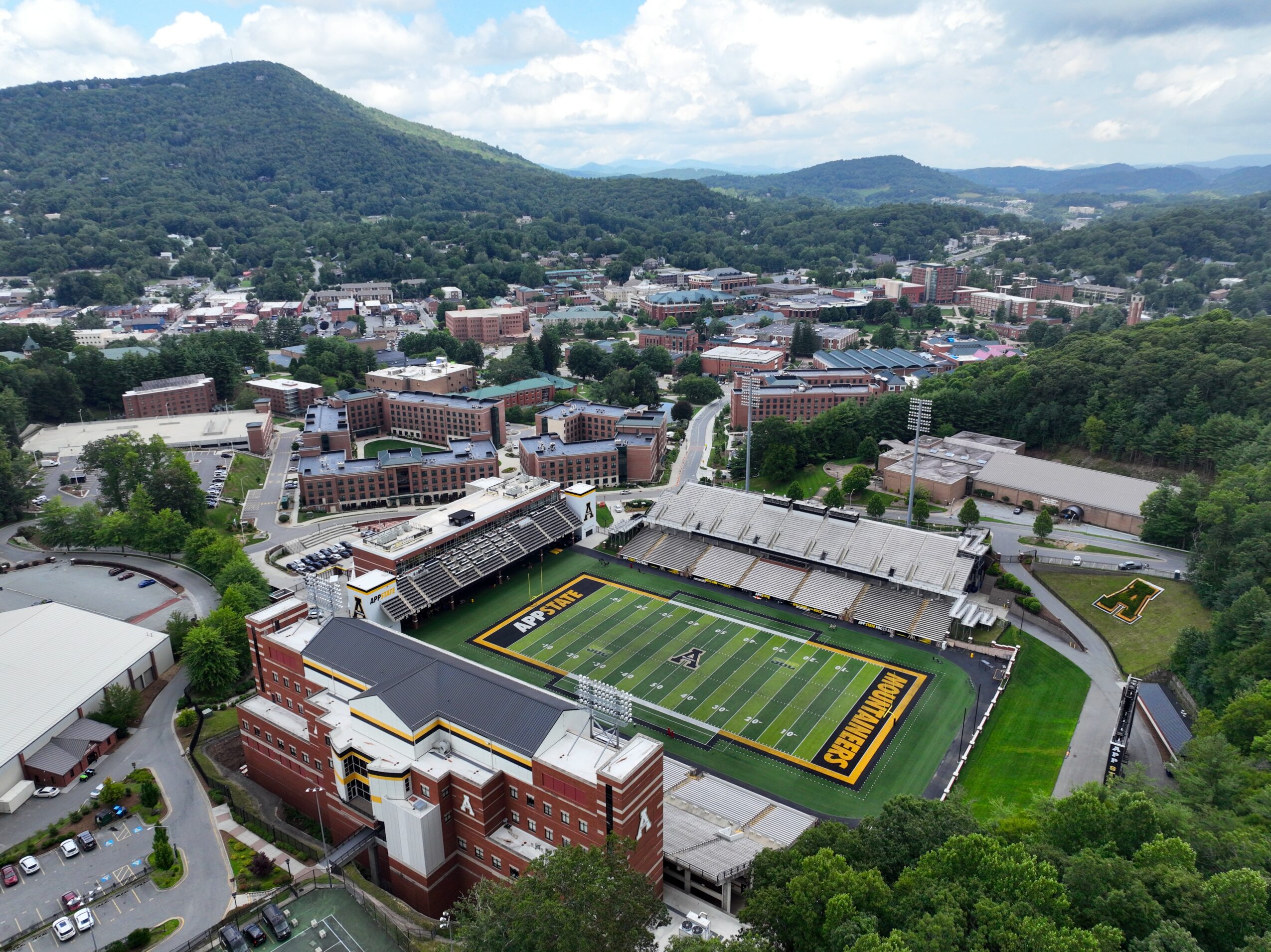 A football stadium with seating and a field, surrounded by buildings and green trees, with mountains in the background.