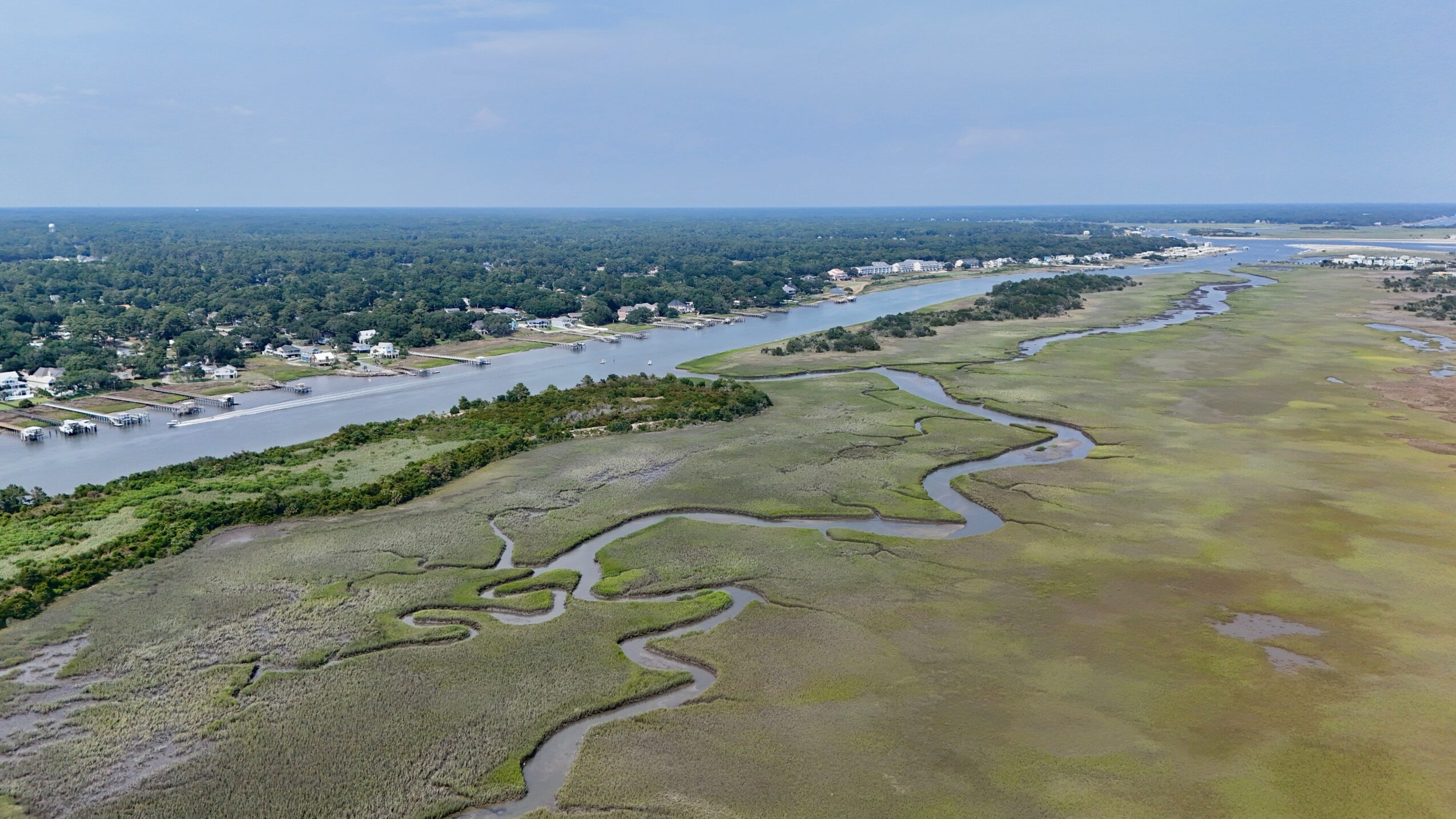 Aerial view of a river winding through a marshland with a distant town and blue sky.