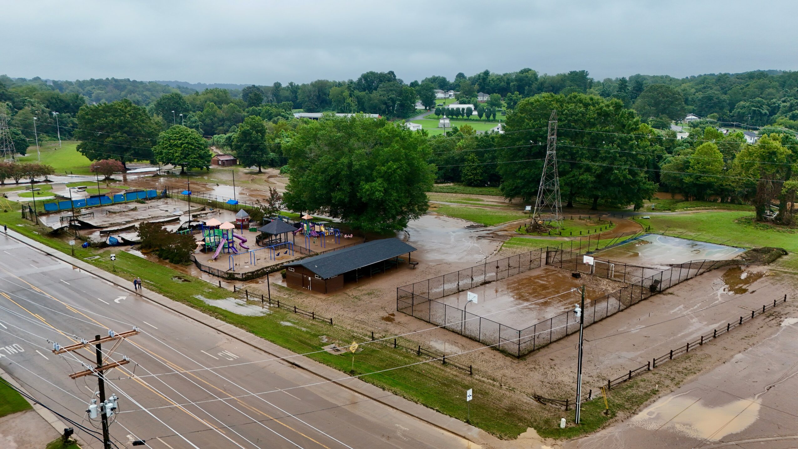 Aerial view of a fenced sports field, nearby buildings, trees, and a road with power lines, under cloudy sky.