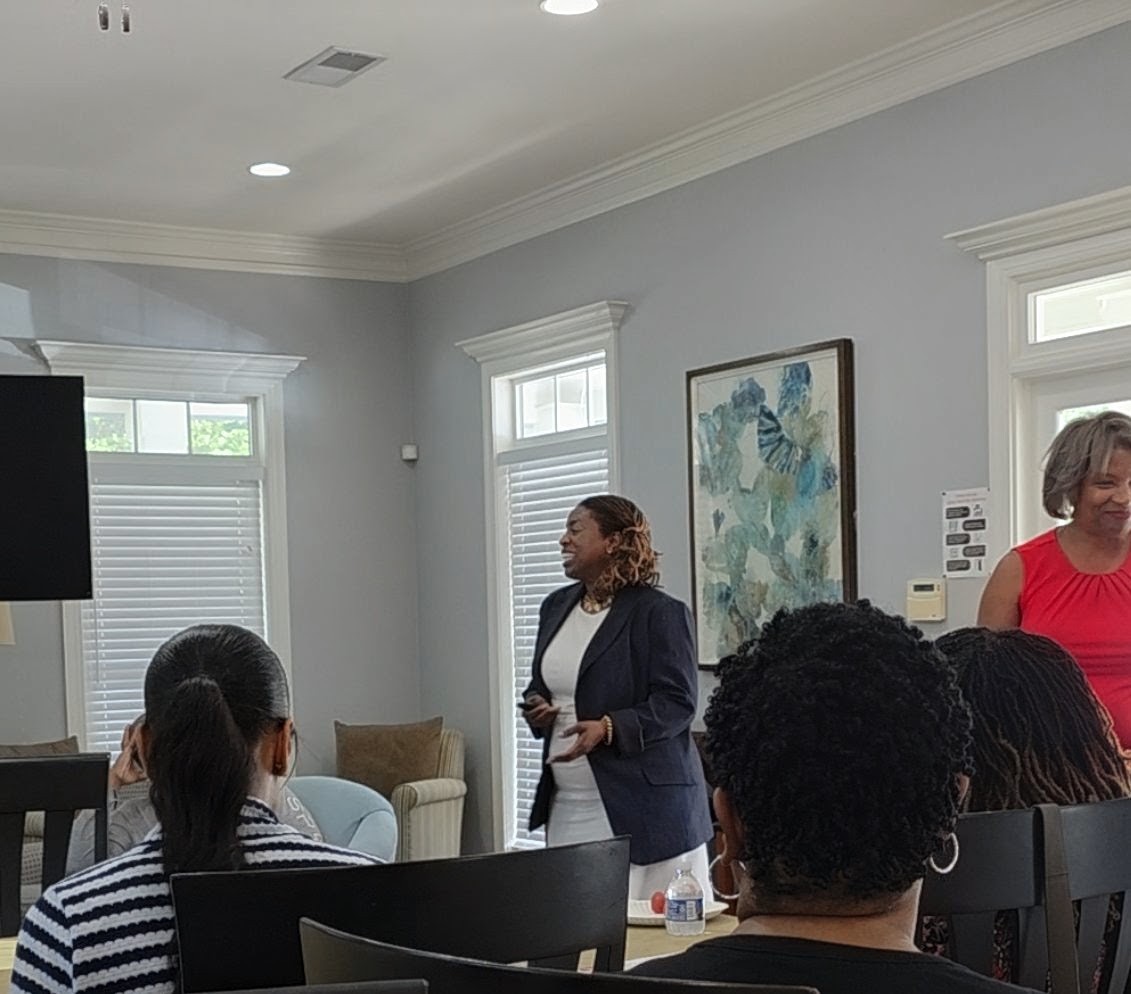 Woman standing and speaking to a group in a bright room with windows and artwork on the wall.
