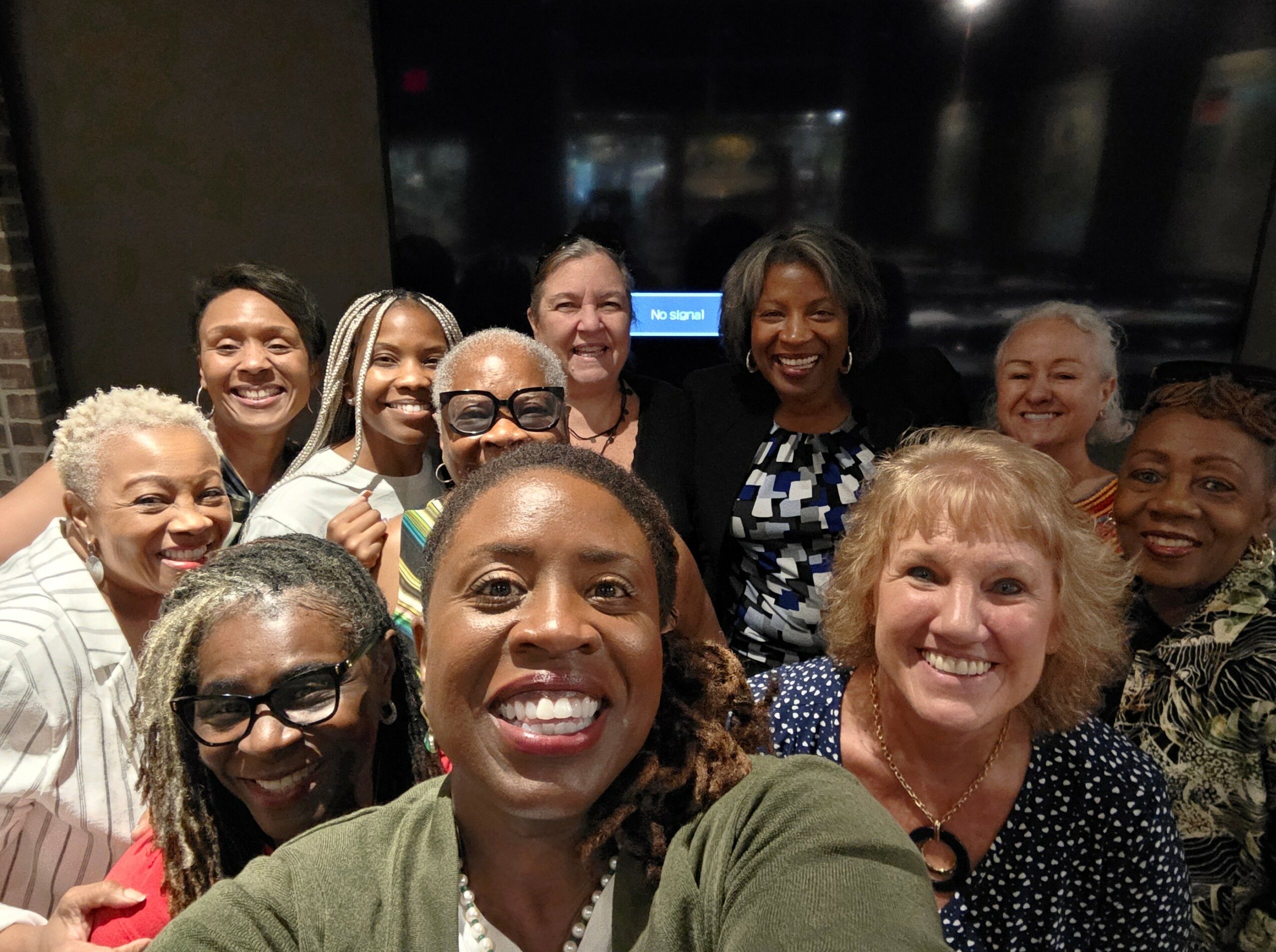 Group of women smiling and taking a selfie indoors, some wearing glasses, diverse in age and appearance.