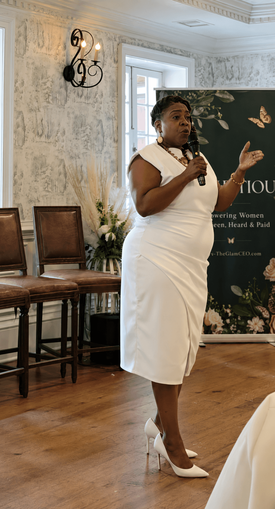 Woman in white dress speaking into microphone in a room with chairs and a floral backdrop.