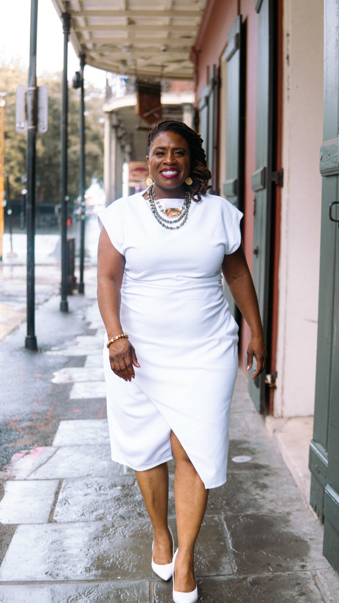 Woman in white dress walking on sidewalk, smiling, holding door handle, city street background, daytime.