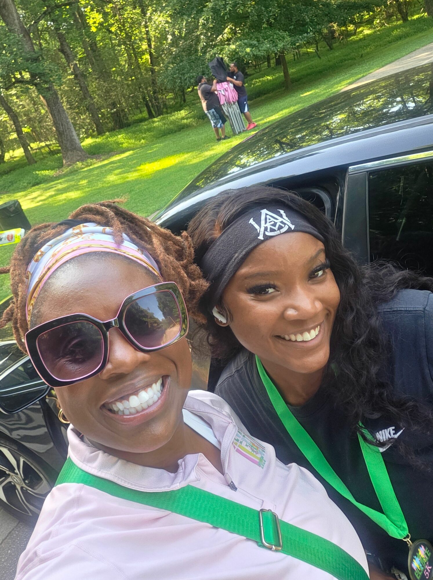 Two smiling women outdoors, one wearing sunglasses and a headband, the other with a badge, near a black vehicle.