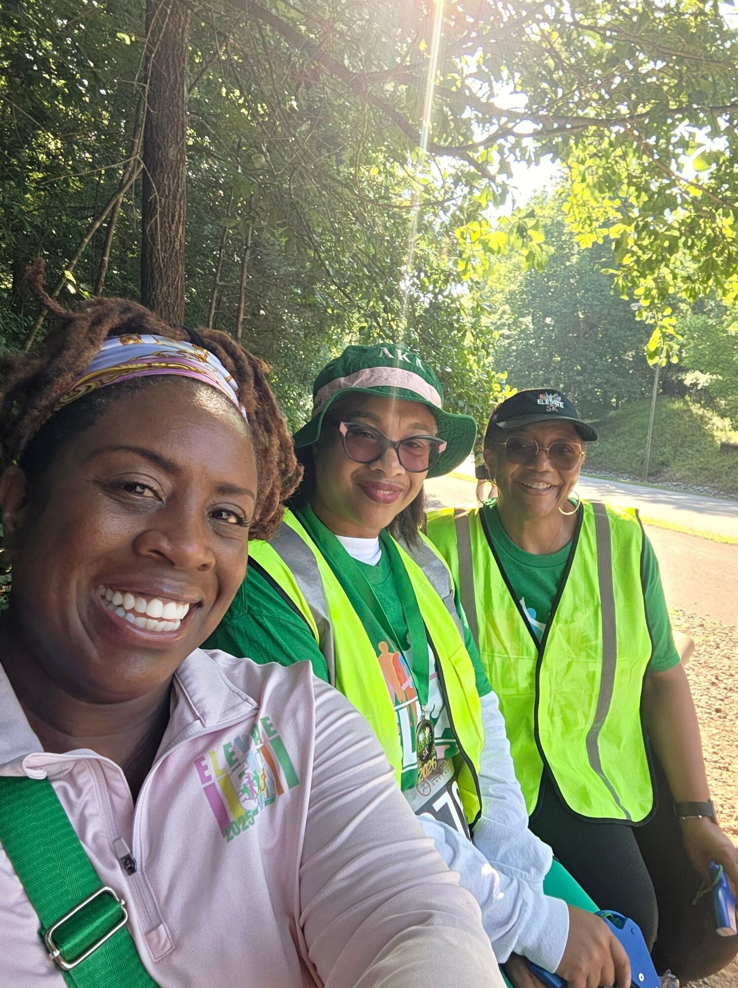 Four people outdoors smiling, surrounded by trees and sunlight, wearing athletic clothing and reflective vests.