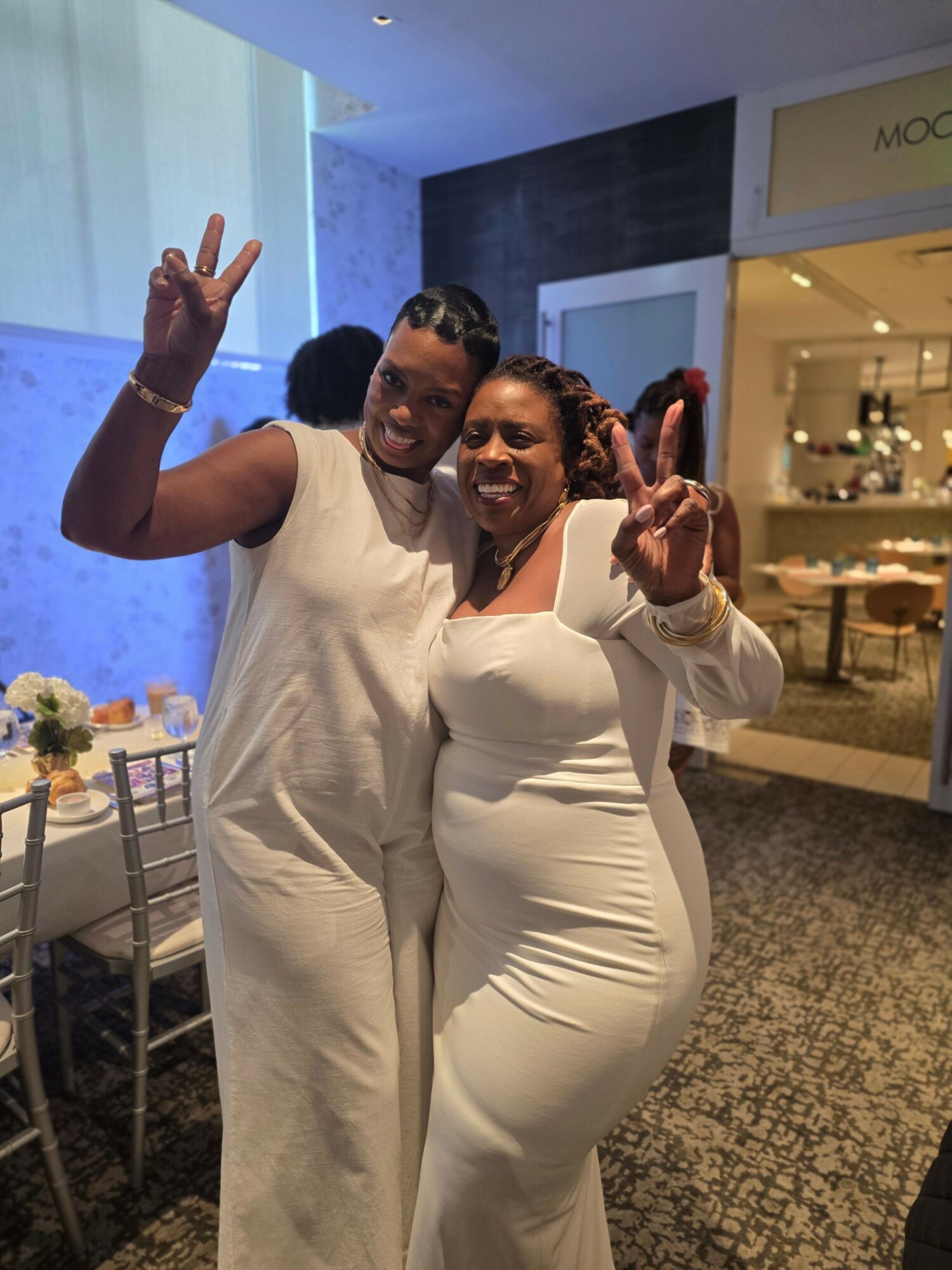 Two women in white dresses smiling and making peace signs indoors, with a table and flowers in the background.