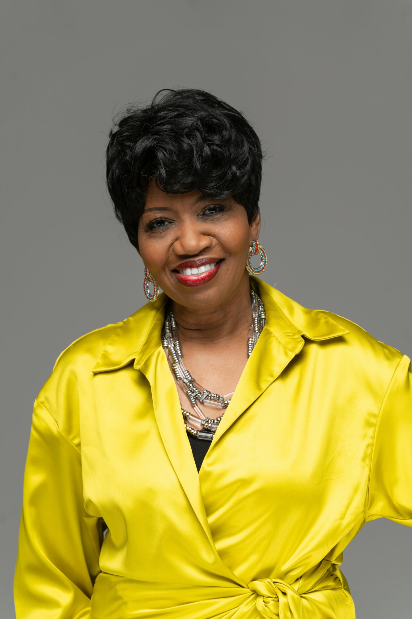 Woman with short curly hair wearing a yellow satin blouse and jewelry, smiling against a gray background.