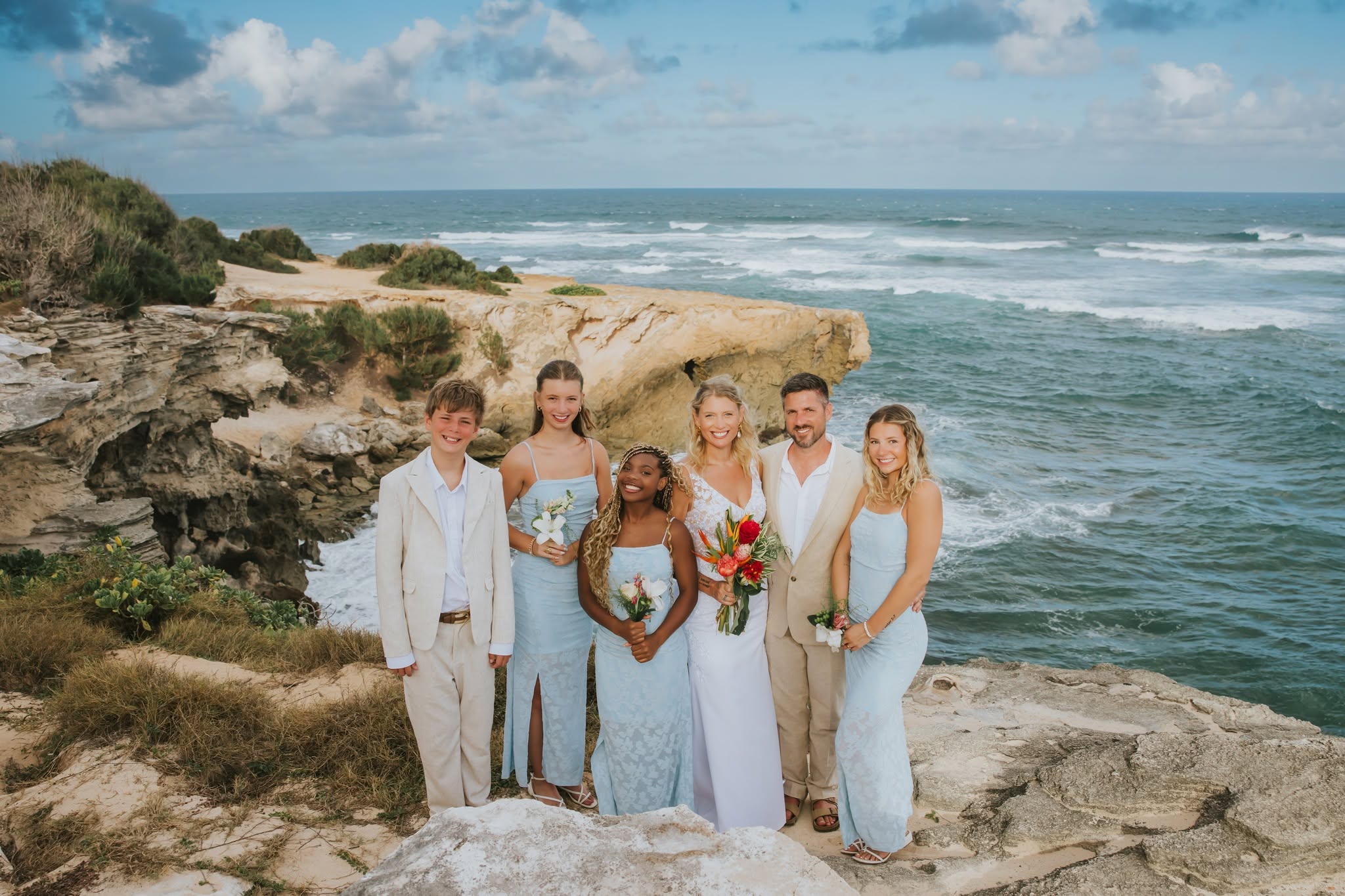 Group of six people, including a bride and groom, standing on a rocky coastline with ocean waves in the background.