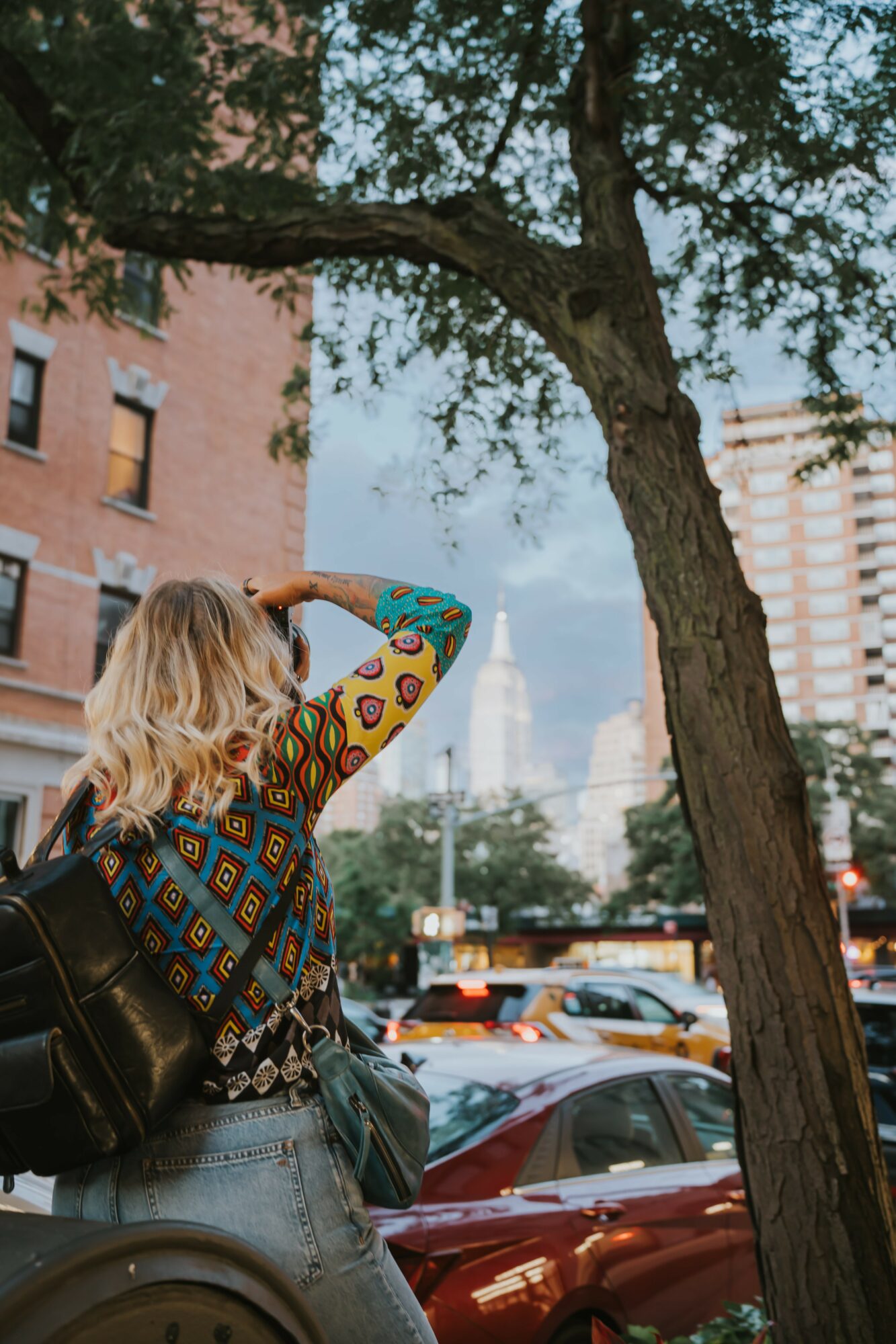 Woman with blonde hair, wearing a colorful patterned top and jeans, standing near a tree in an urban setting with buildings and cars.