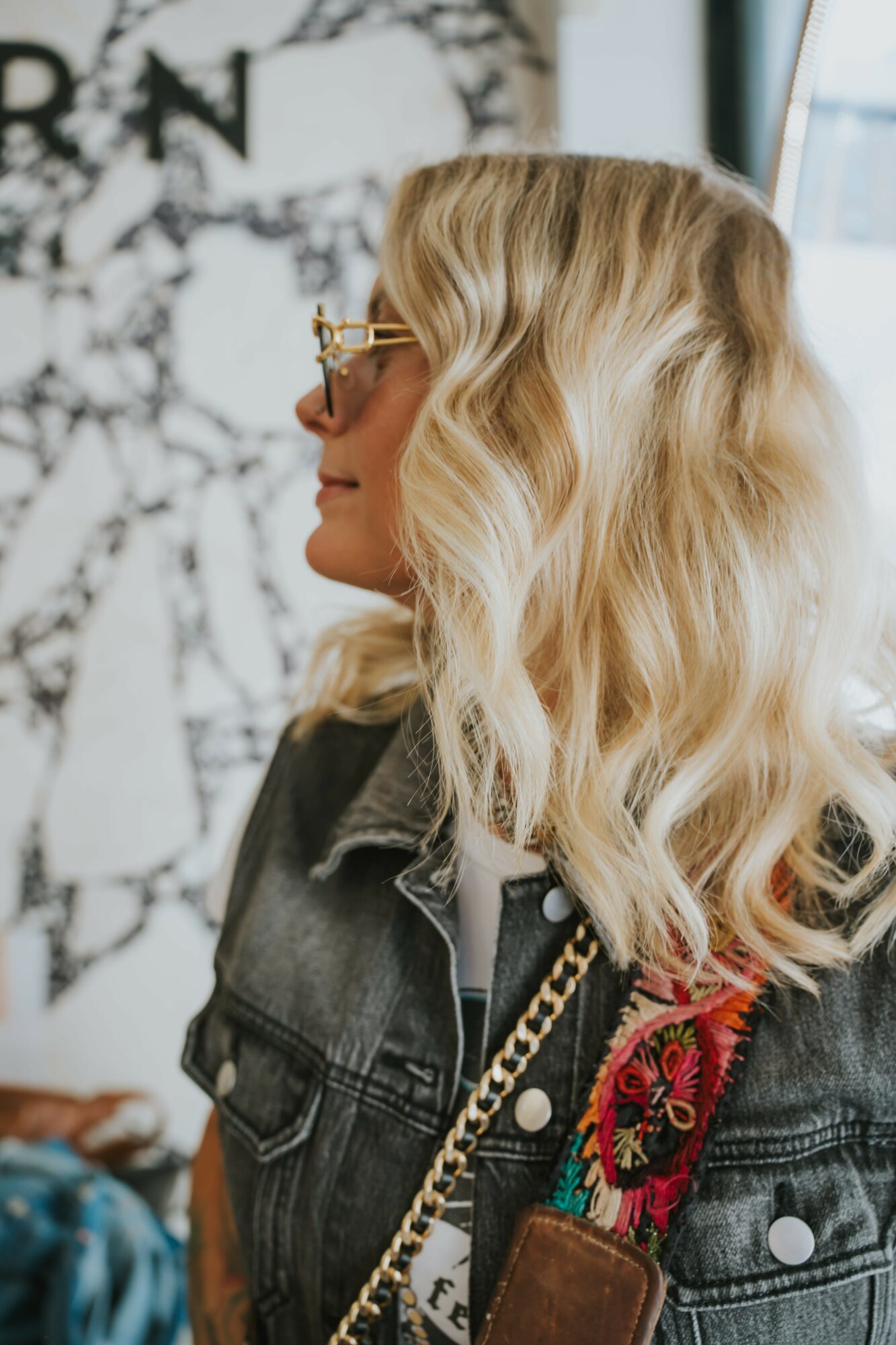 Side profile of a woman with wavy blonde hair, wearing glasses and a denim jacket, indoors near a window.