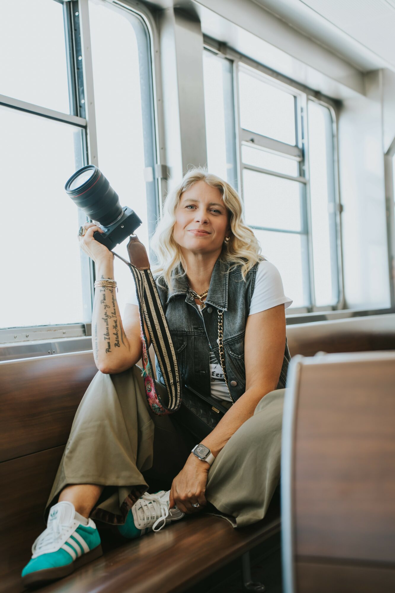 Woman sitting on a bench holding a camera, inside a building with large windows, smiling.