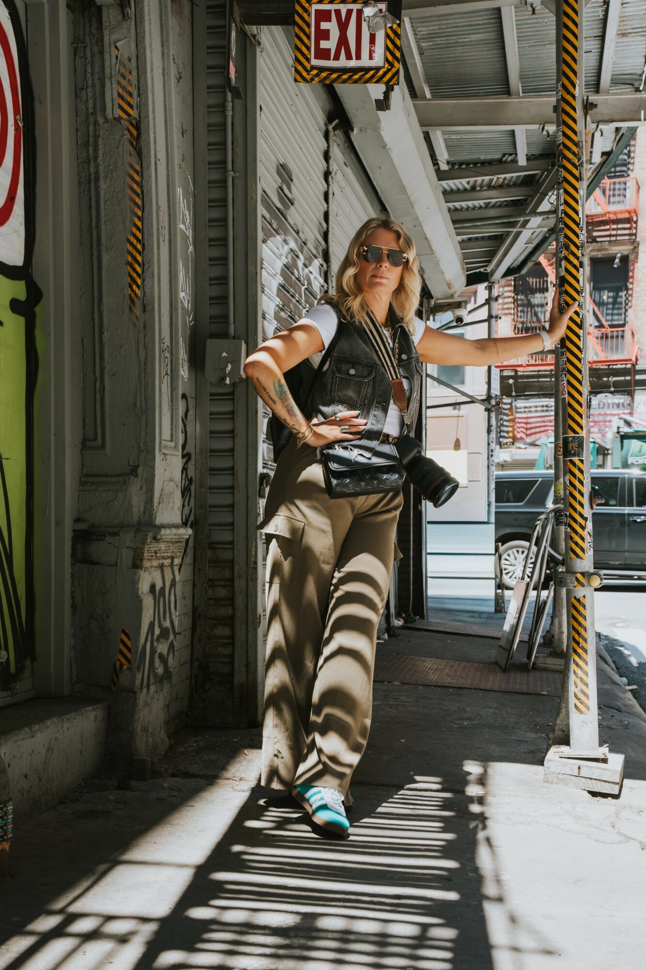 Woman with sunglasses and a leather vest stands in a sunlit urban alleyway, leaning against a wall with shadows cast by overhead structures.