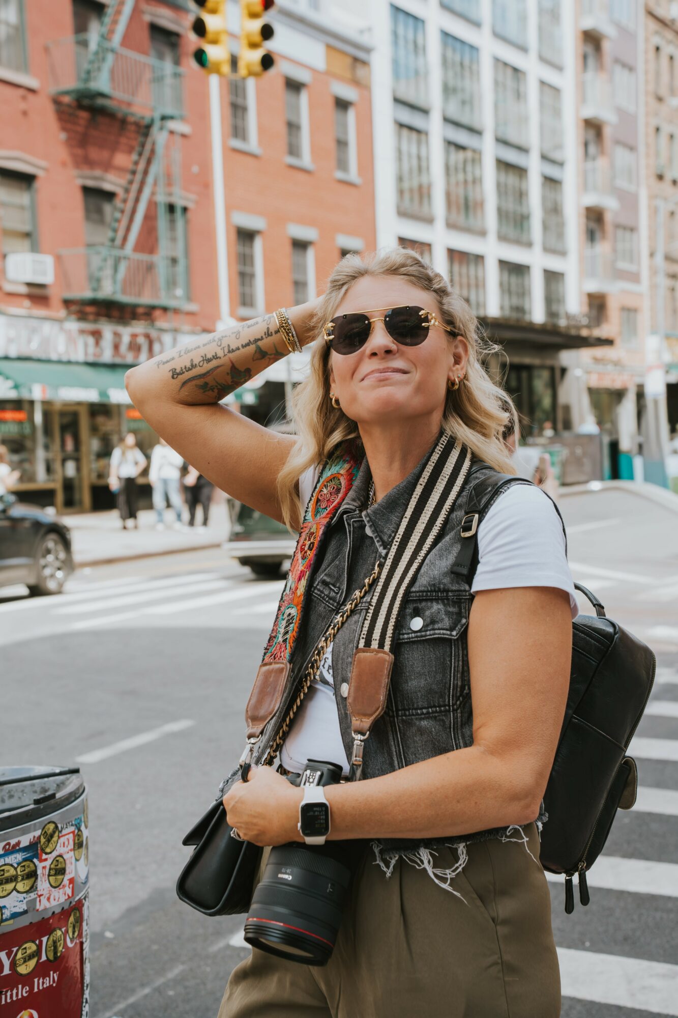 Woman with sunglasses and a backpack standing on city street, smiling, with buildings and pedestrians in background.