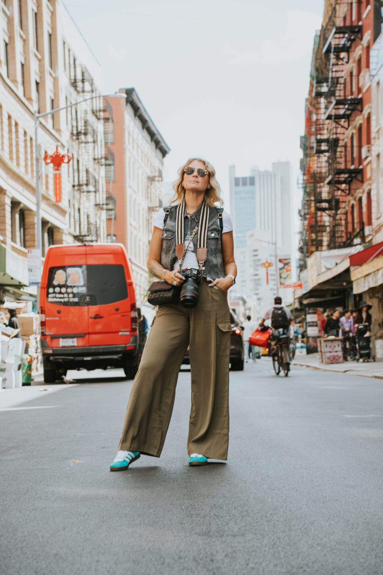 Woman standing in city street with buildings, vehicles, and pedestrians, holding a camera and wearing sunglasses, vest, and wide pants.