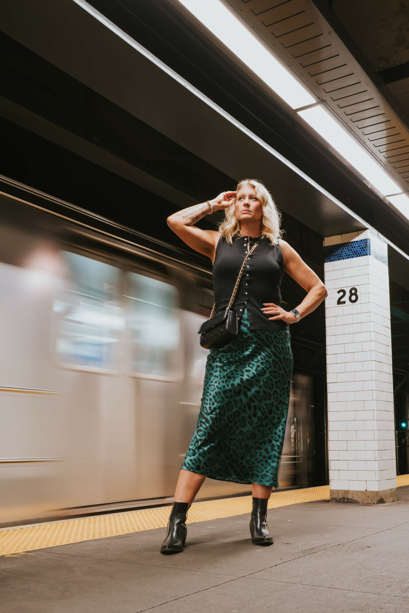 Woman with blonde hair in a green skirt, black top, and boots stands on subway platform, looking to the side.