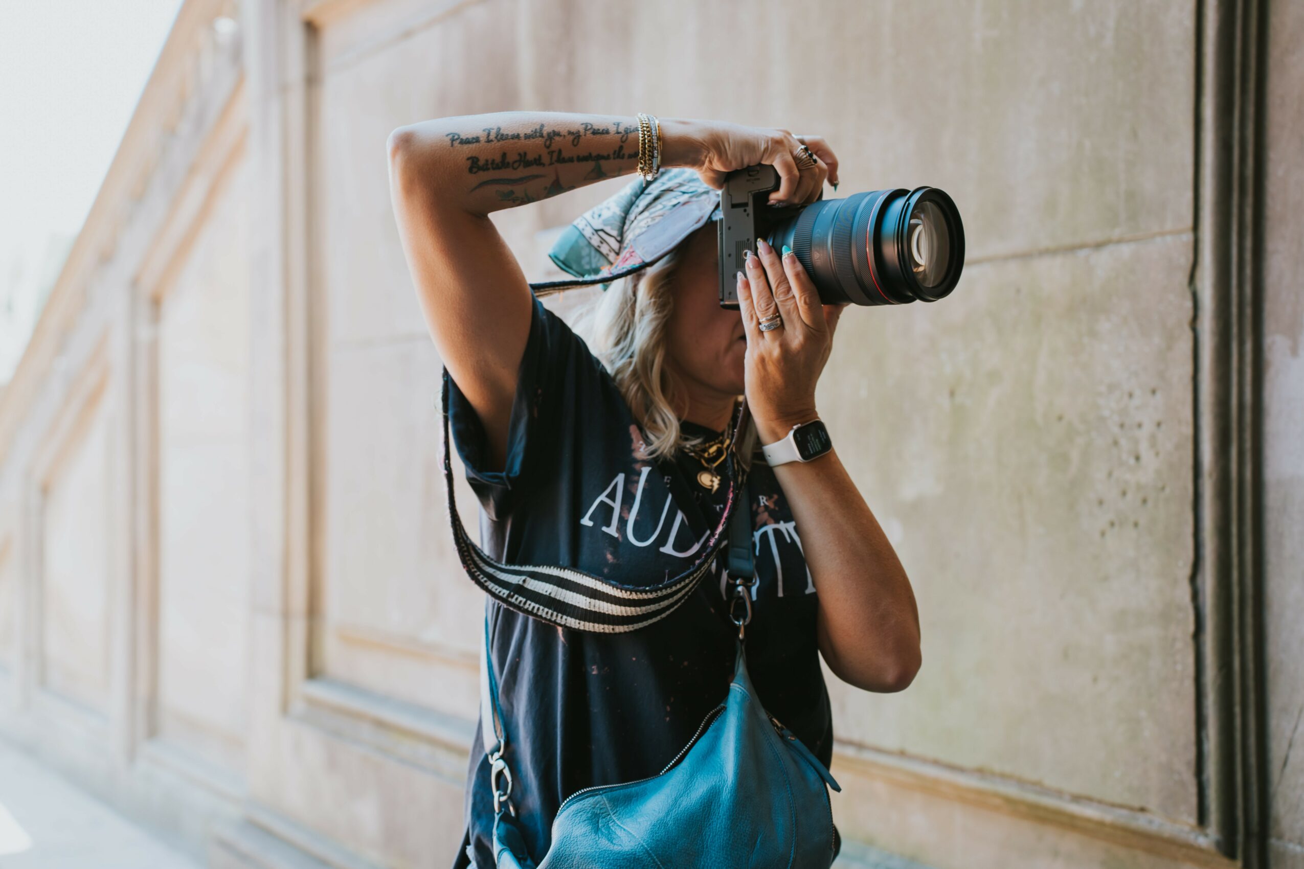 Person holding a camera with a large lens, wearing a black and blue sports jersey, standing against a stone wall.
