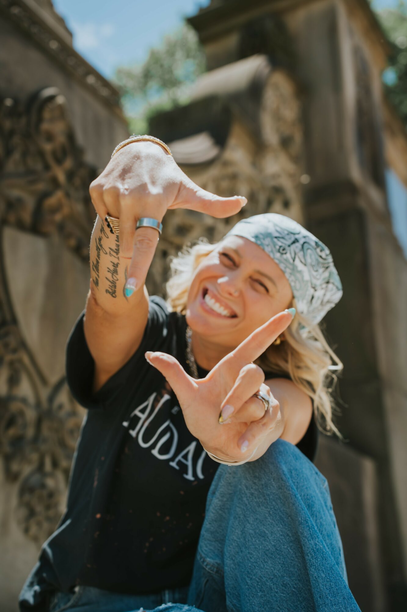 Smiling woman with a bandana, pointing at a soap bubble outdoors, with a stone structure in background.