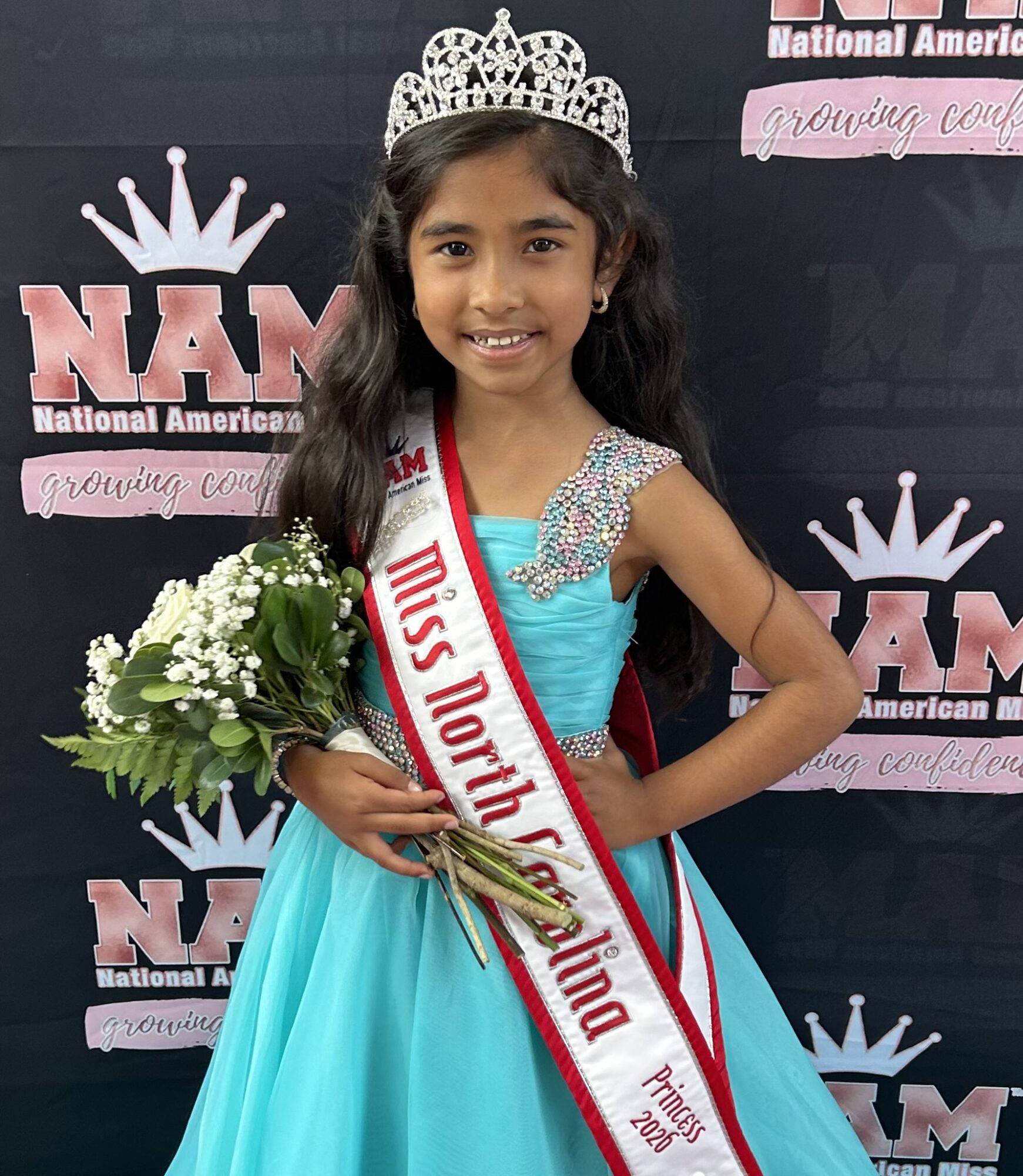 Young girl wearing a crown and sash, holding flowers, smiling, in front of a backdrop with logos and text.