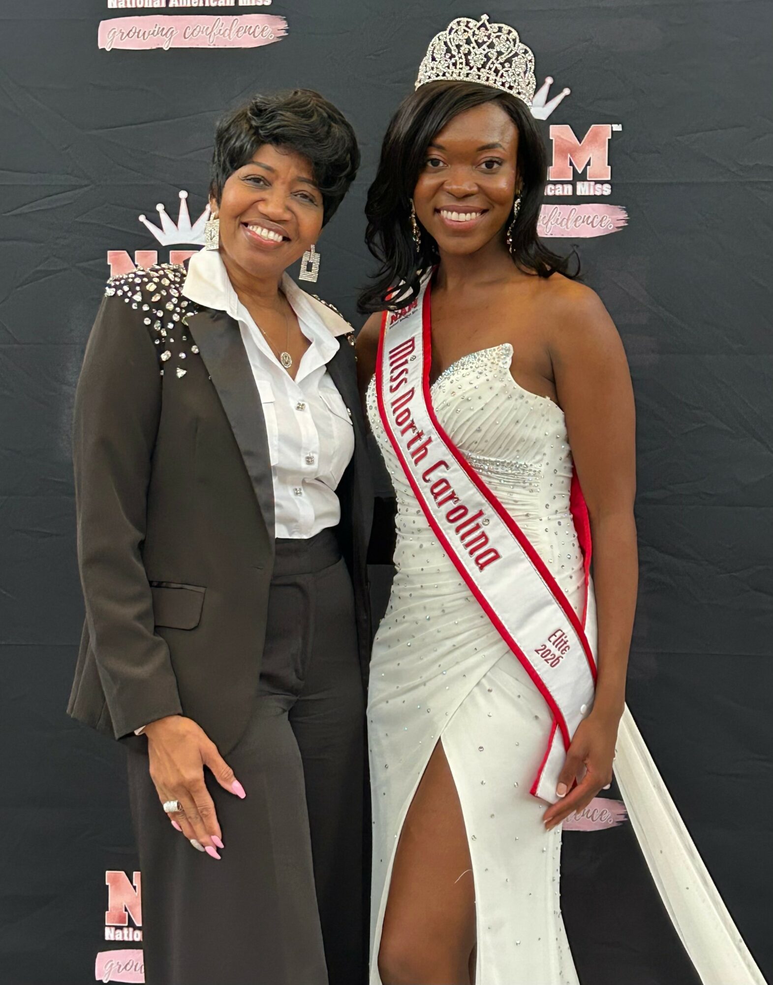 Two women smiling, one wearing a crown and sash, the other in a suit with embellishments, standing in front of a black backdrop.