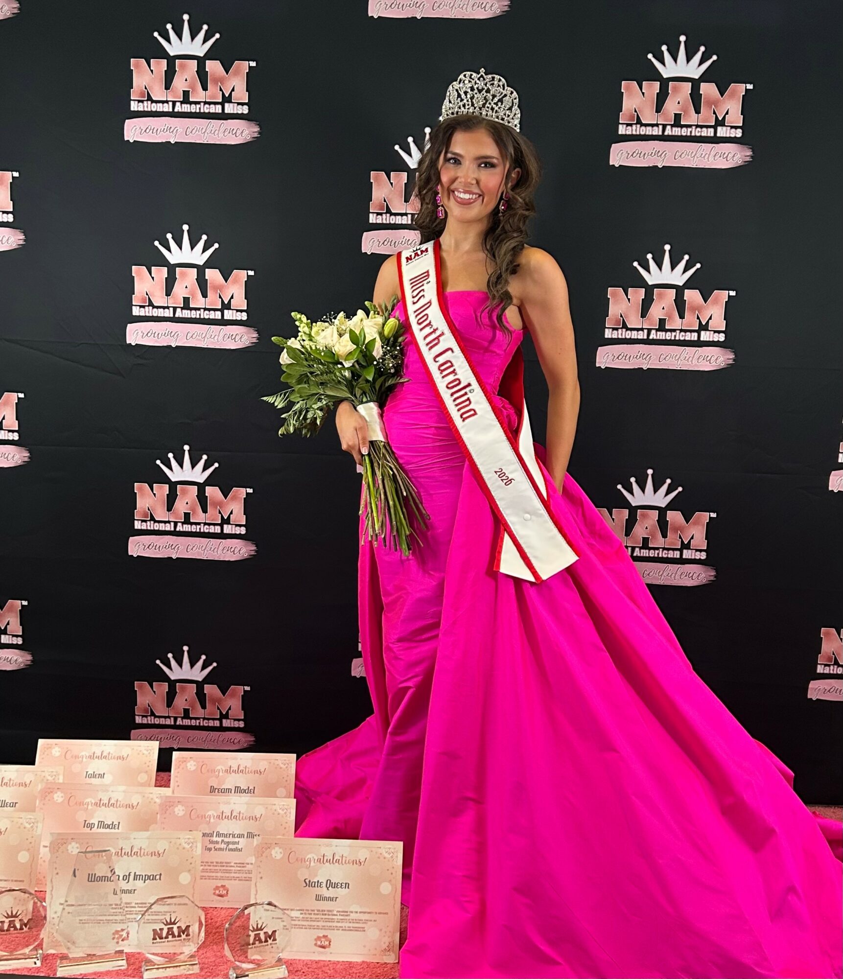 Woman in pink gown with crown and sash holding flowers, standing in front of NAM backdrop, smiling.