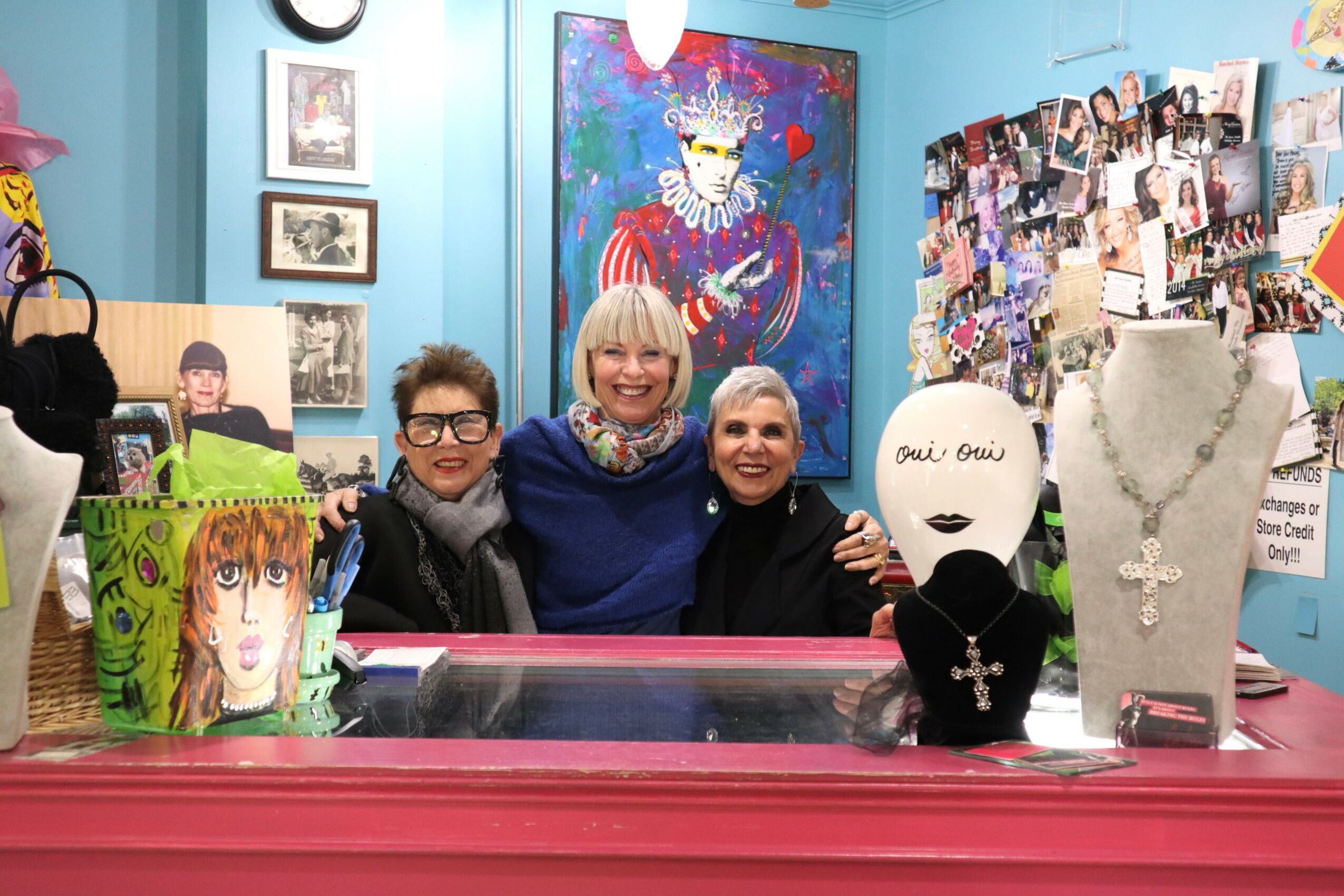 Three women smiling behind a pink display counter in an art gallery, with artwork and photos on the walls.