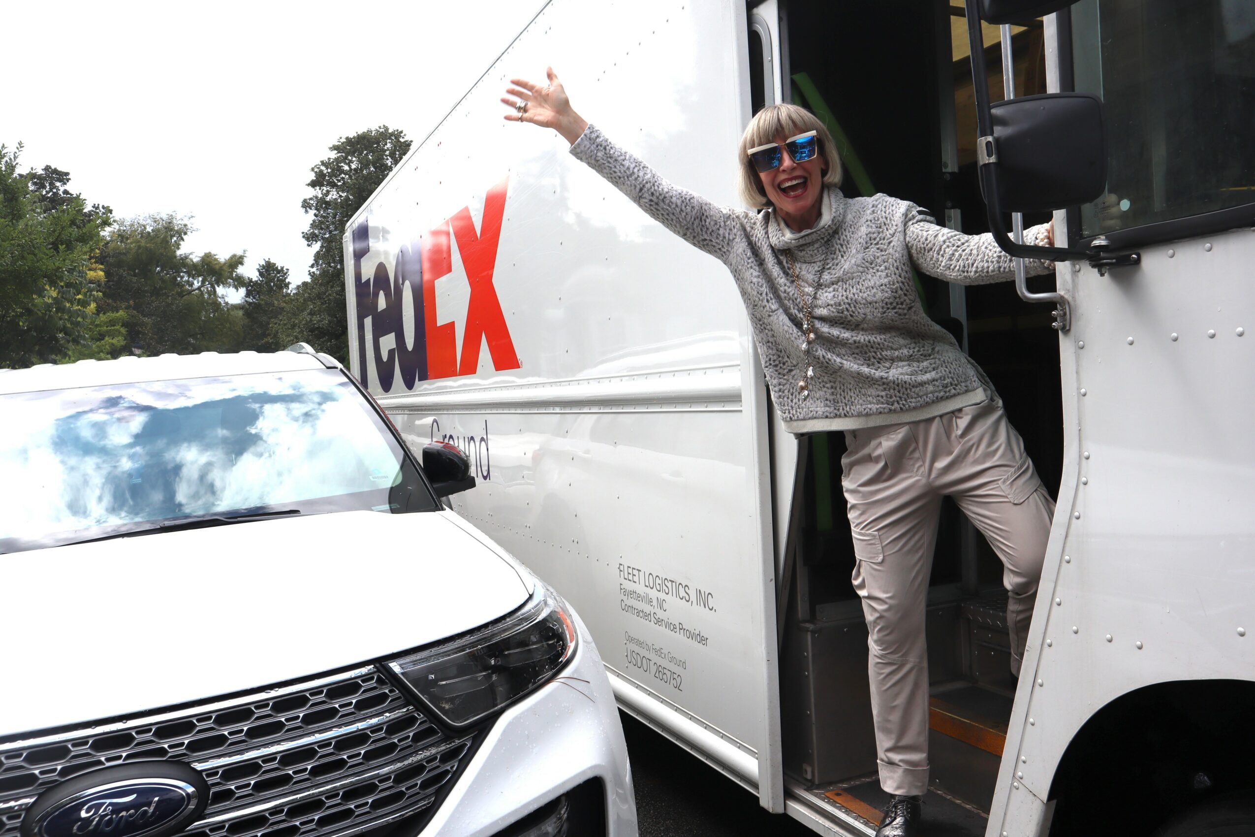 Person with sunglasses and gray outfit standing in FedEx truck doorway, smiling and waving outside, with a white car nearby.