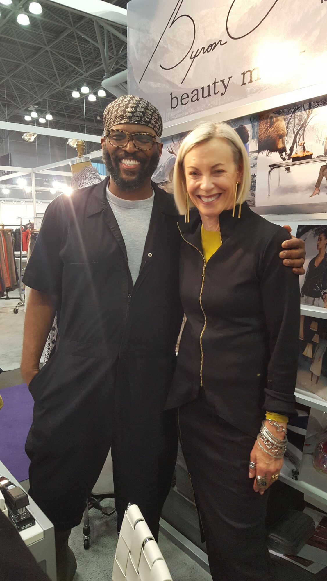 Two people standing together indoors, one wearing a black suit and the other in a black jacket with a yellow shirt.
