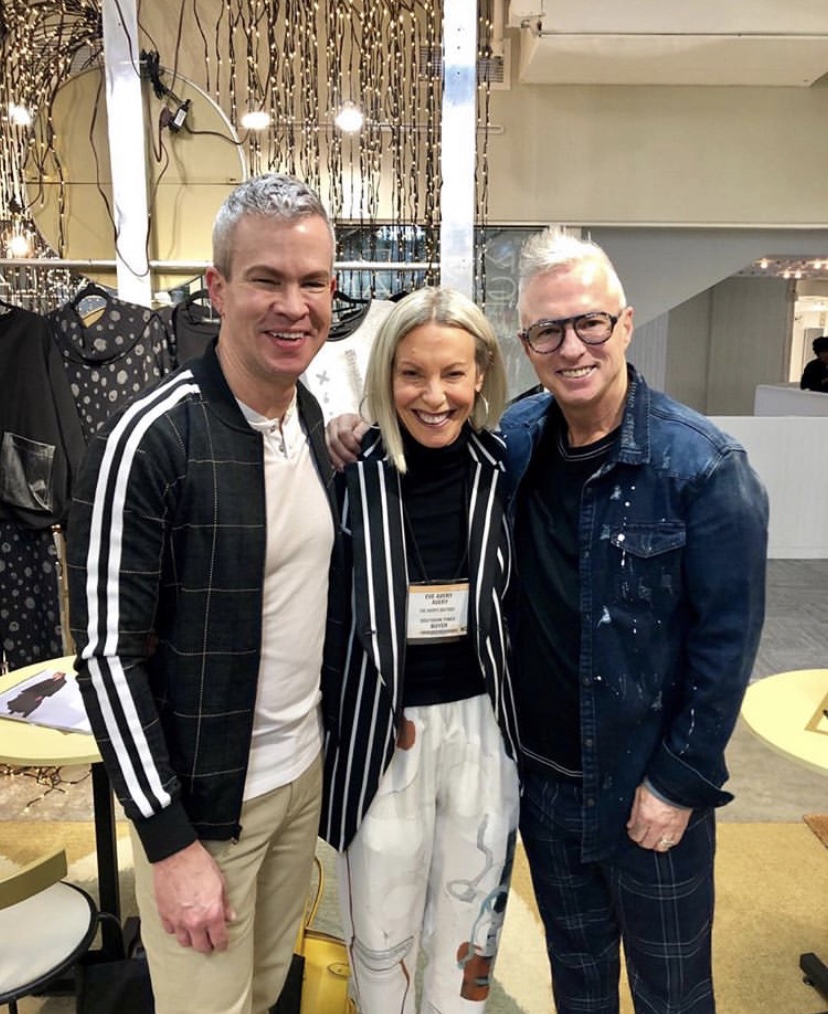 Three people smiling indoors, two men and one woman, standing close together, with a background of curtains and furniture.