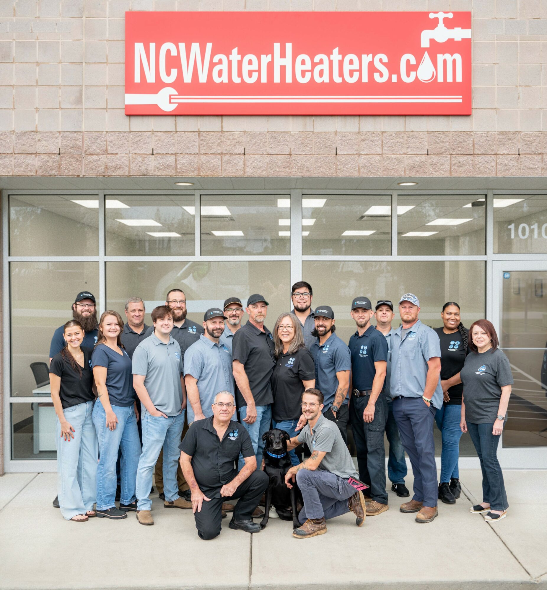 Group of people standing outside a store with a sign reading NCWaterHeaters.com, in front of a glass storefront.