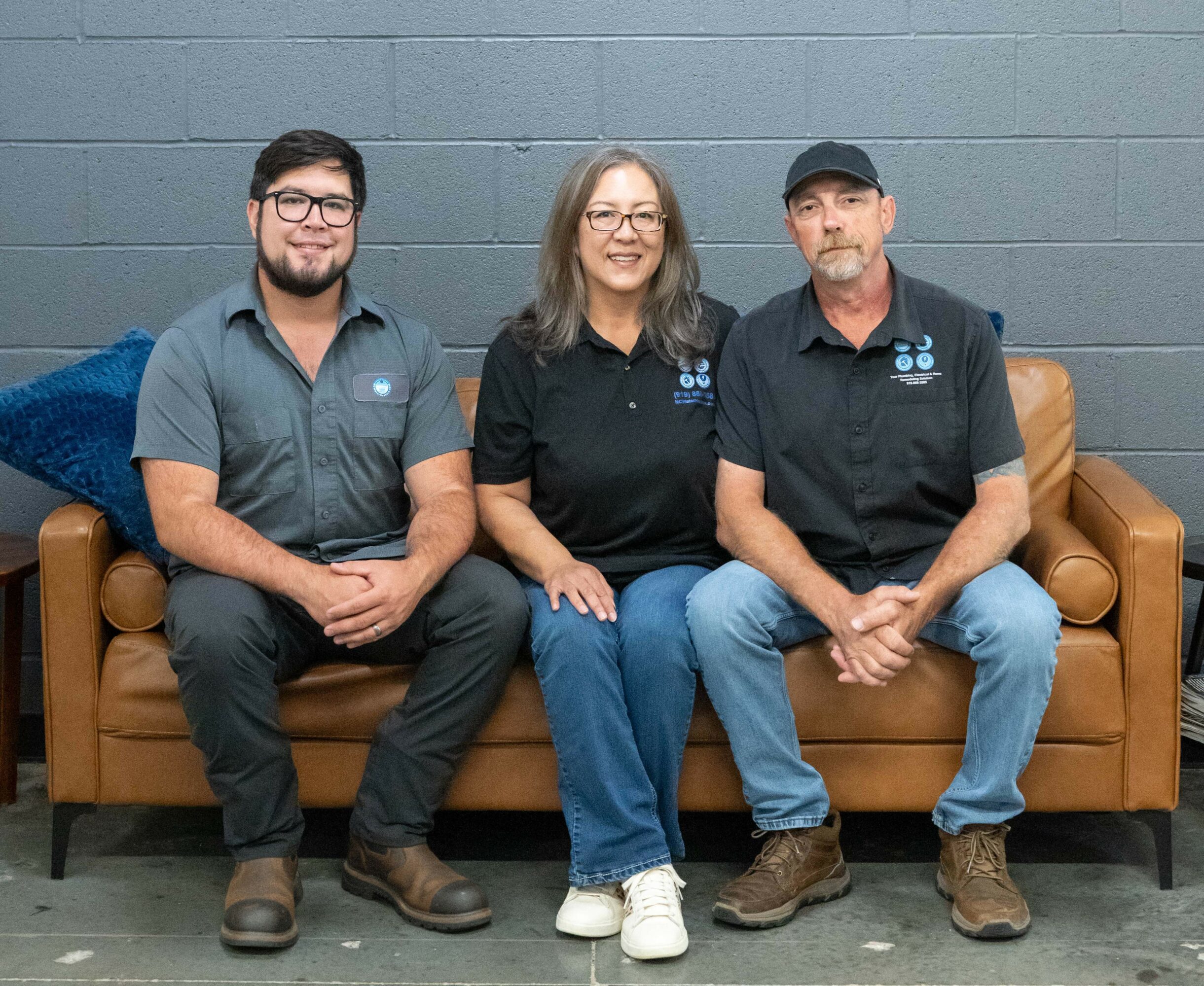 Three people sitting on a brown couch against a gray wall, smiling, with a blue pillow on the left side.