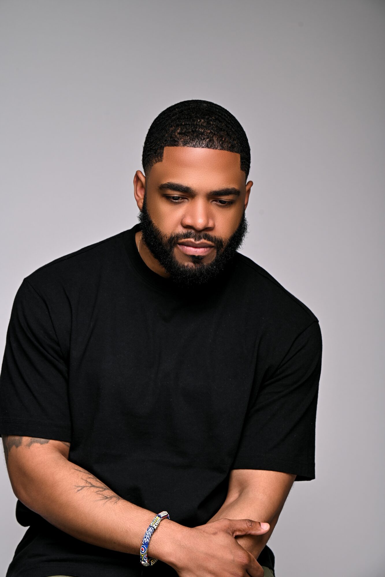 Man with short hair and beard wearing a black shirt, looking down, arms crossed, neutral expression.