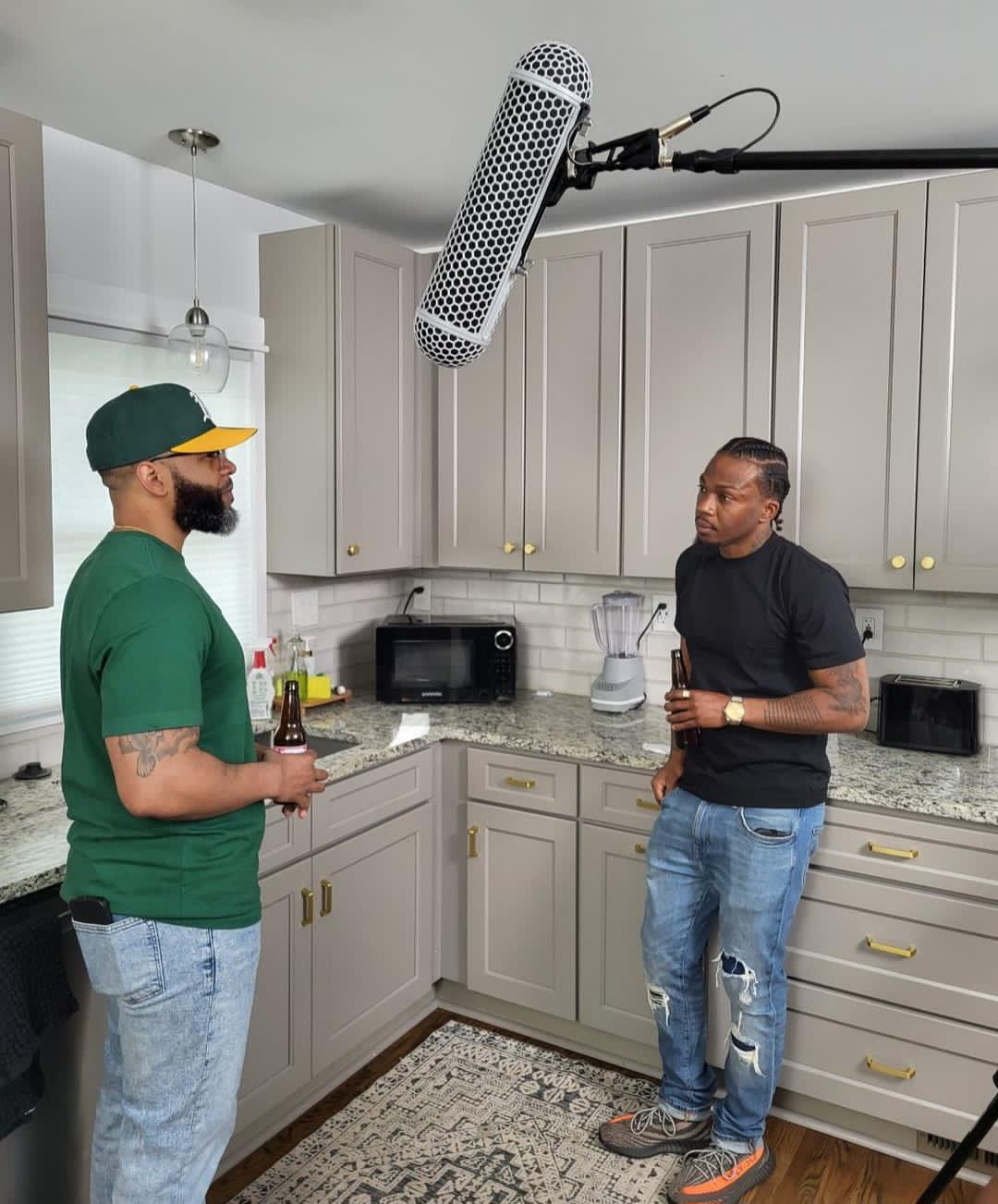 Two men stand in a kitchen, holding drinks and talking, with a microphone hanging above them. Kitchen has white cabinets and a patterned rug.
