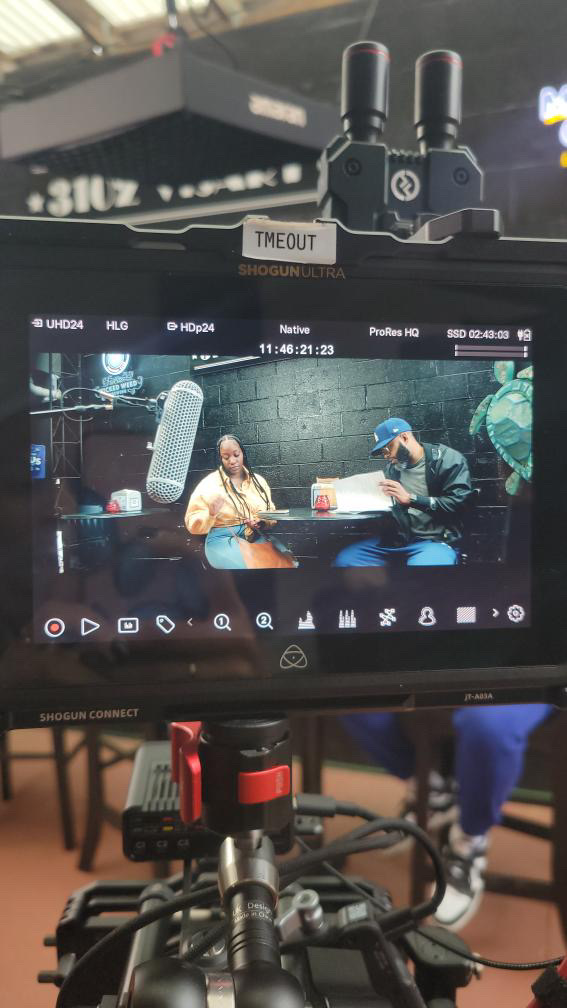 Camera monitor showing two people sitting at a table, with equipment and a person in a blue cap nearby.