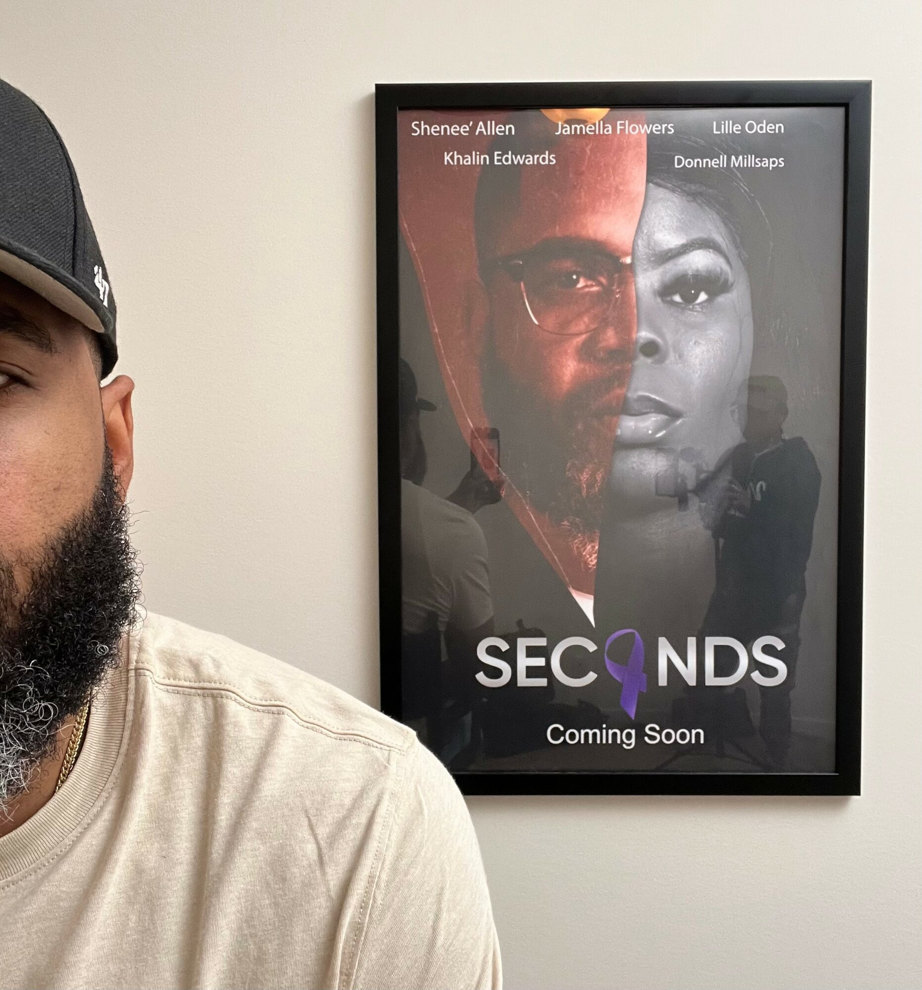 Part of a person's face and shoulder visible on the left, with a framed poster on the wall behind showing two faces and text.