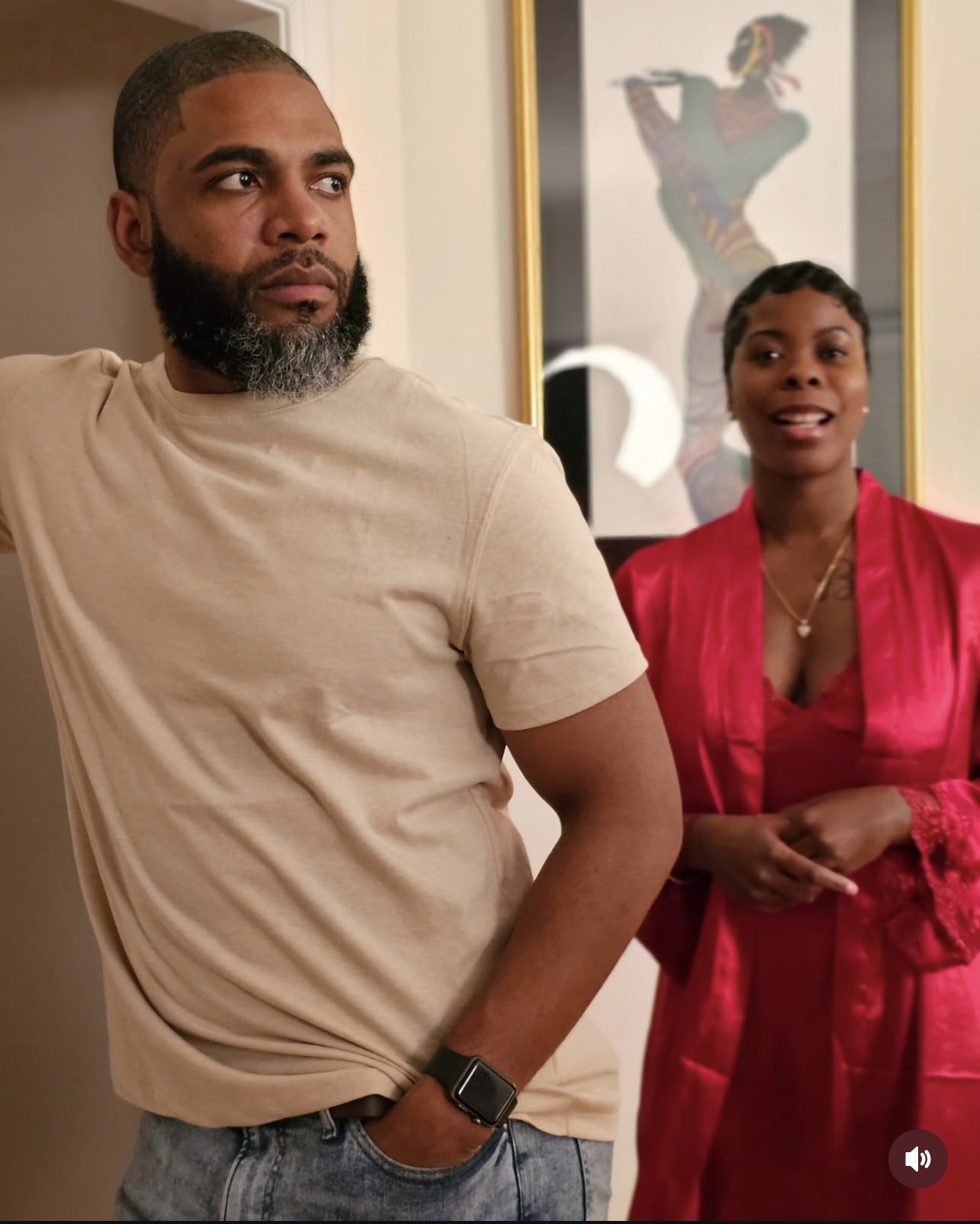 A man with a beard and a woman in a red dress stand indoors, with a framed picture of a woman in the background.